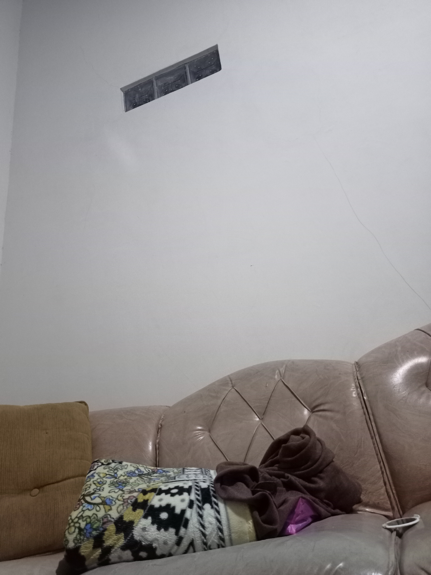 The image shows an indoor scene, likely a living room, with a plain white wall and a portion of a tufted leather couch. On the couch are several objects: a light brown corduroy pillow on the left, a patterned blanket or throw in the center, and a dark brown folded piece of fabric on top of the blanket. A small section of a window made of translucent blocks is visible high on the wall. The environment appears to be dimly lit, and there are faint lines or cracks visible on the wall. There are no people or animals present in the image. The location of Karangtalun, Indonesia is not ascertainable from the visual information provided in the image.