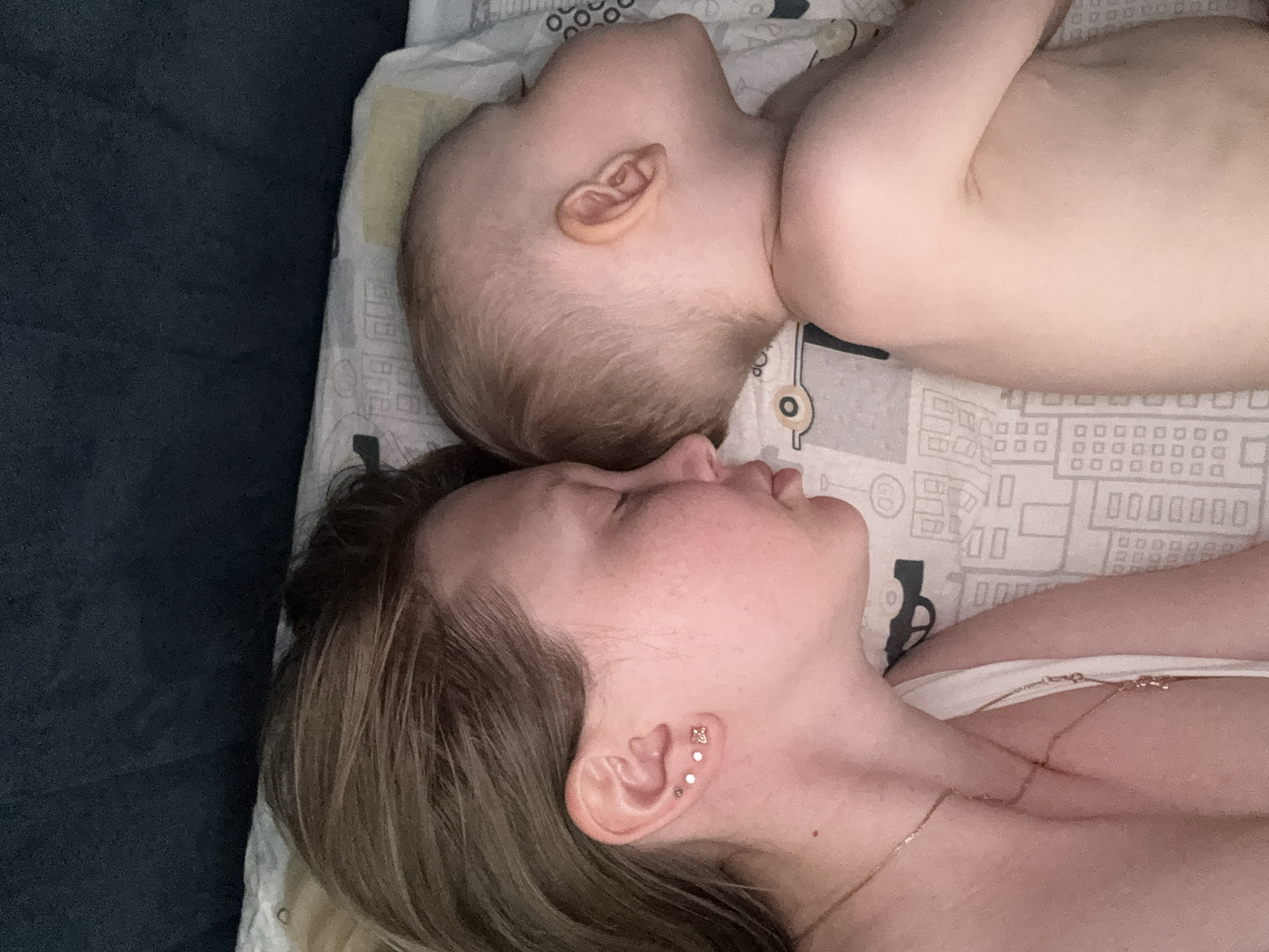 A close-up, overhead shot shows a woman and a baby lying down, side-by-side, on a patterned surface. The baby is positioned above the woman, with its head tilted back and facing upwards. Its ear is clearly visible, with fine blonde hair around it. The baby's body is mostly out of frame, but its smooth, pale skin is visible.
The woman is lying below the baby, her head tilted towards the baby's. Her eyes are closed, and her mouth is slightly parted. She has blonde hair that falls across her forehead and shoulder. Her left ear is visible, adorned with multiple small earrings, including a flower-shaped one. A delicate necklace is visible on her collarbone and draped across her chest. She is wearing white undergarments. The background is a pale fabric with a repeating pattern of buildings and geometric shapes in grey and black. A dark, textured surface is visible on the left side of the frame, possibly a piece of furniture. The lighting suggests it is daytime. The overall mood is intimate and peaceful, implying a moment of rest or sleep shared between mother and child. There is no visible text in the image.