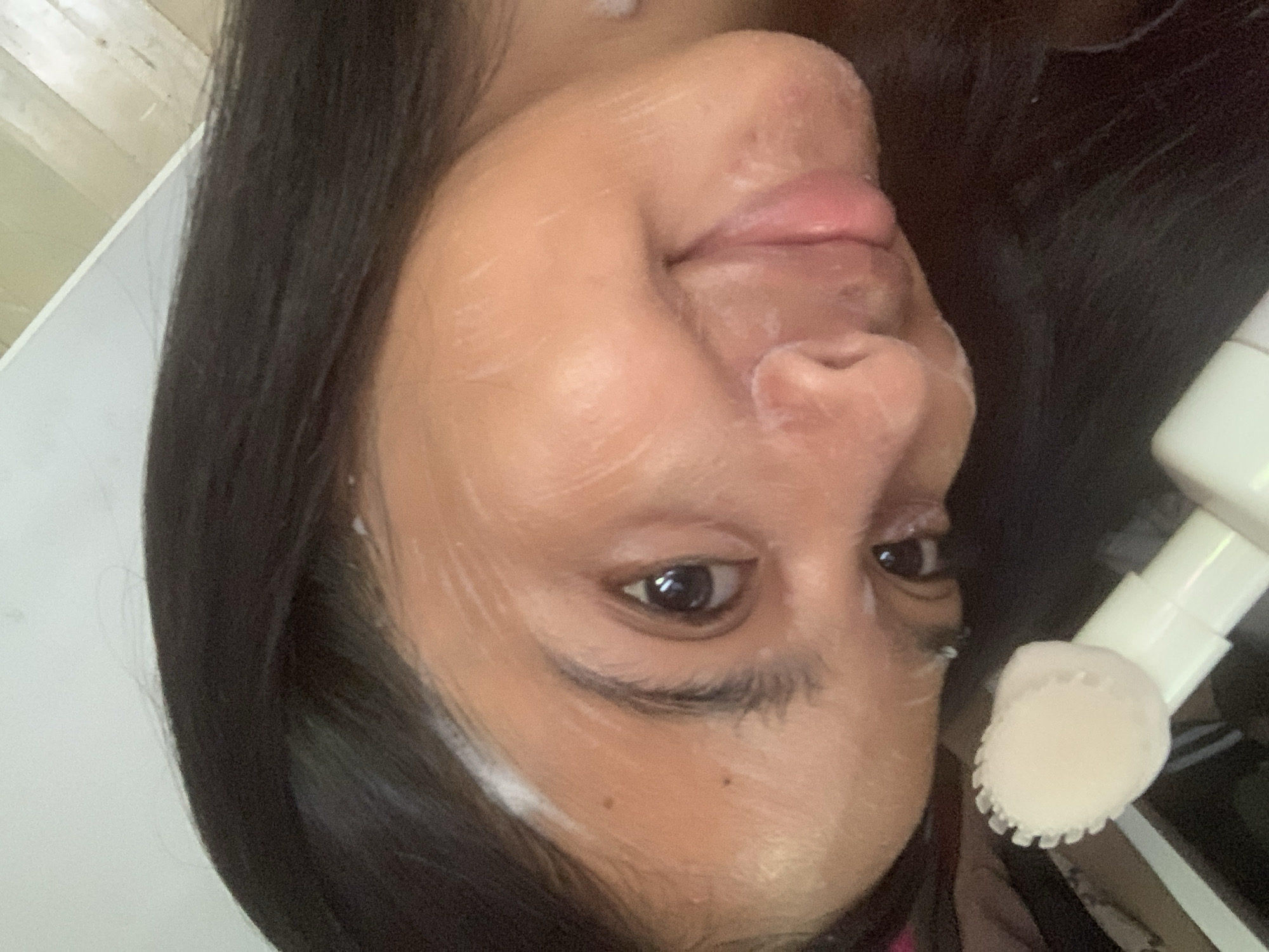 The image, captured in Sungai Meriam, Indonesia, presents a close-up of a person's face, slightly tilted, in an indoor setting. The individual has dark hair and medium-toned skin. Their face is largely covered with a light-colored, creamy substance, suggesting the application of a facial cleanser or mask.
The person's eyes are open and looking directly at the camera, conveying a calm or slightly smiling expression. Their lips are slightly parted. On the right side of the frame, a white facial cleansing brush, featuring a soft, textured head, is visible near the person's cheek, indicating a skincare routine is in progress. The creamy substance is evenly spread across the face, avoiding the immediate eye area. Notable details include two small moles on the right cheek below the eye.
The background is simple and light-colored, suggesting an interior space with ambient lighting. There is no visible text in the image.