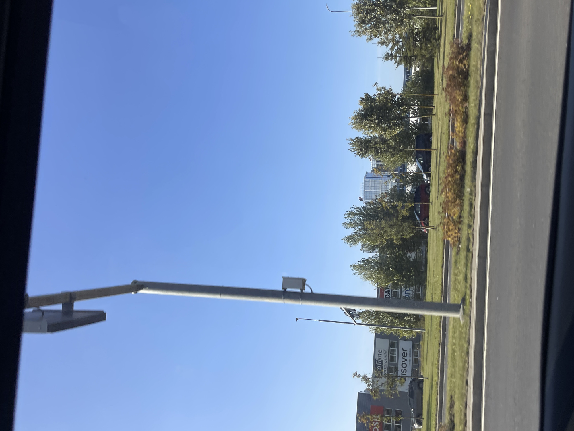 The image shows a street view with buildings and trees under a clear blue sky in Tallinn, Estonia. In the foreground, a road is visible with a median strip of grass and bushes. A tall pole with a box attached to it stands prominently on the right side of the image. In the background, several buildings are present, including one with the text "ProfiLine" and "isover" visible on its facade. Trees line the area between the road and the buildings. A few cars are parked or driving on the road. The overall scene suggests an urban or suburban commercial area.