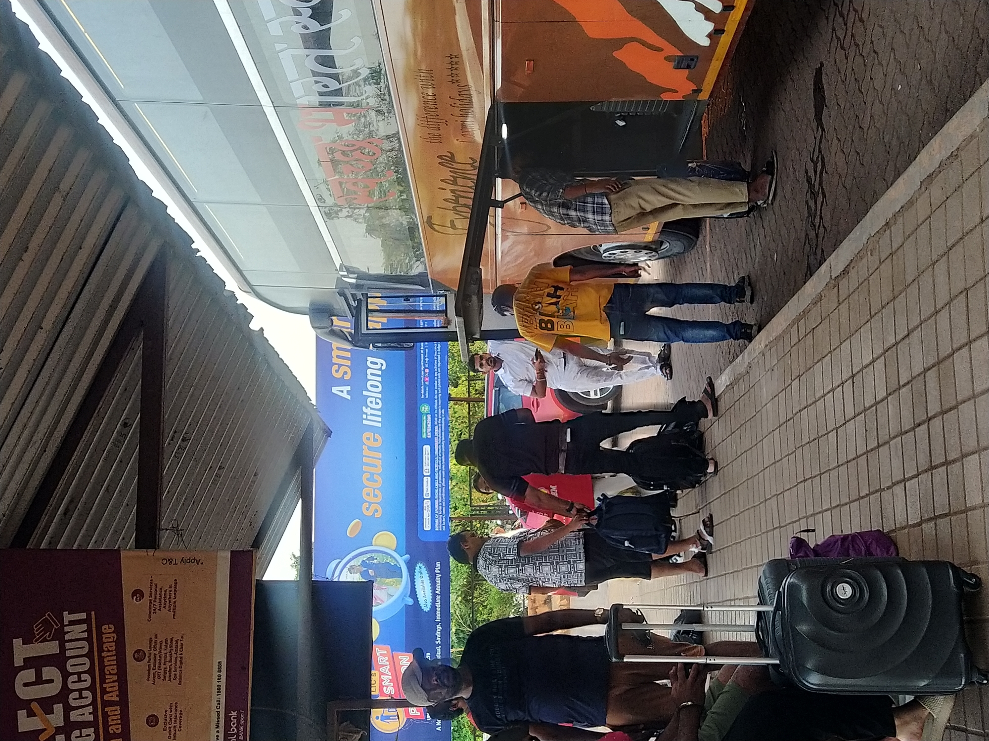 The image shows a bus station in Mapusa, India. A large, orange and silver bus is parked at the curb. Several people are standing or walking near the bus, some with luggage. They appear to be boarding or disembarking. Advertisements for financial services, including LIC's SMART PLAN and a bank account, are visible on a nearby structure. The area is covered by a roof. The ground is paved with interlocking bricks.