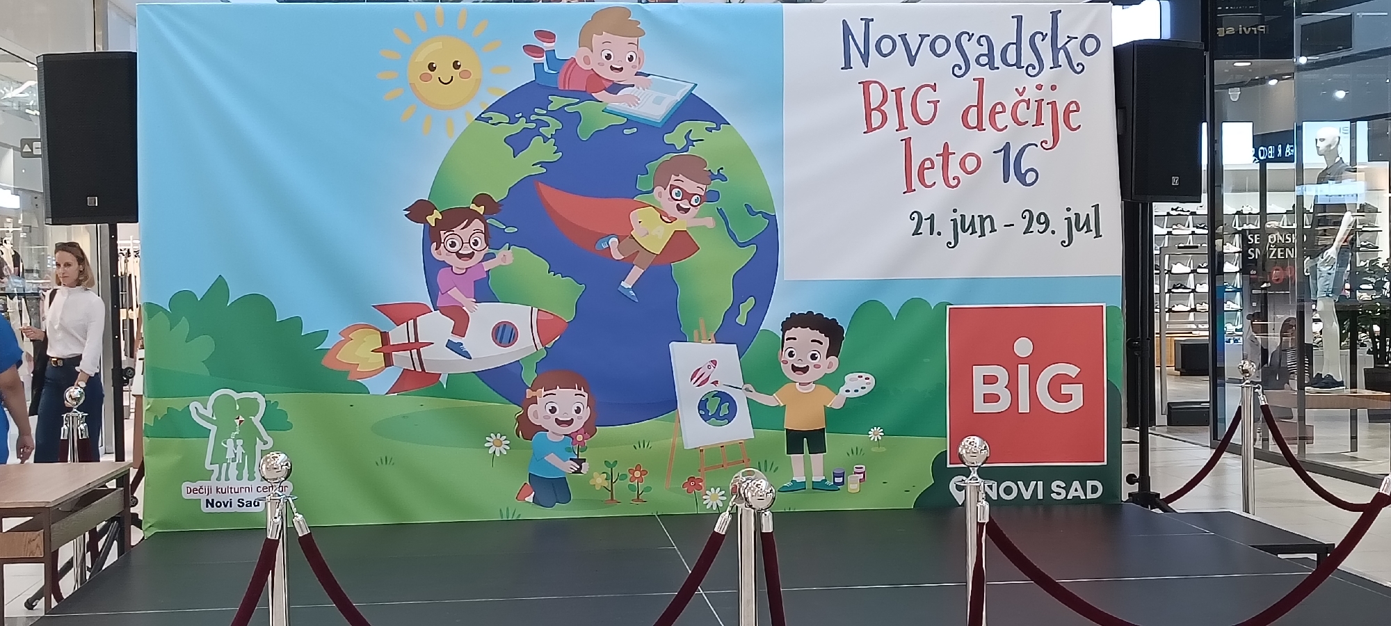 This media file shows a large banner depicting a vibrant, cartoon-like scene with children and elements related to space exploration and art. The banner is set up in what appears to be an indoor public space, possibly a mall or event venue, indicated by the presence of retail displays and mannequins visible in the background through a glass partition. Red velvet ropes and stanchions are set up in front of the banner and a stage, suggesting an event is either about to take place or has recently concluded.
The banner itself features a cartoon Earth with several children engaged in activities. One child is reading a book on top of the Earth, another is flying like a superhero with a cape, and a third is riding a rocket ship. Below, a girl is planting flowers, and a boy is painting a picture that includes the Earth and a rocket. The overall aesthetic is bright, cheerful, and child-friendly, with a sunny sky and green grassy hills.
Text on the banner includes "Novosadsko BIG dečije leto 16" which translates to "Novi Sad BIG Children's Summer 16," along with the dates "21. jun - 29. jul" (June 21 - July 29). There is also a logo for "BIG" and text indicating "Dečiji kulturni cenar Novi Sad," meaning "Children's Cultural Center Novi Sad."
A woman in a white shirt and dark pants is partially visible to the left of the banner, near a large speaker. The lighting suggests it is daytime. The scene conveys a sense of summer, fun, and creativity aimed at children.