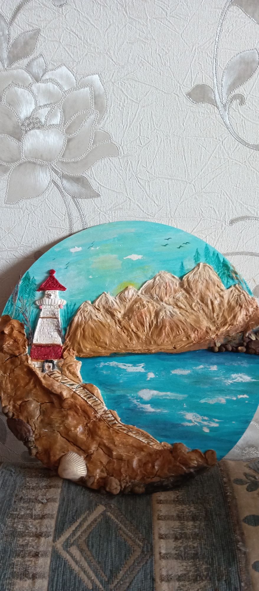 A round artwork featuring a lighthouse, mountains, and water is visible. The lighthouse is white with a red roof and base, perched on a brown rocky cliff. A staircase leads from the lighthouse down the cliff towards the water. Behind the cliff are brown and tan mountains under a blue sky with sparse clouds and birds. The water is blue with white highlights. The artwork is placed against a wall with a floral pattern. A seashell is attached to the cliff. The artwork is situated in an unspecified location within the city of Unknown, Unknown.