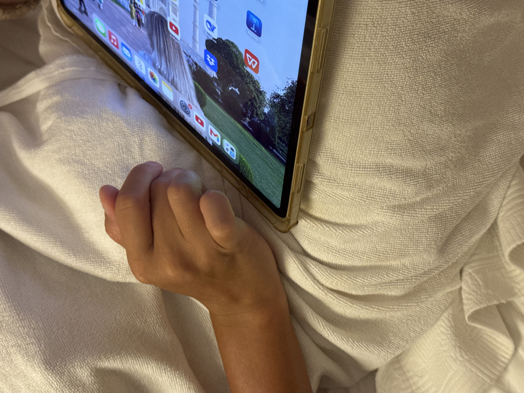 A close-up, eye-level shot shows a person's arm and hand resting on a textured, off-white blanket. The hand is resting on the blanket, with the palm facing upwards. To the right of the arm and hand, a tablet is partially visible. The tablet screen displays a grid of app icons, with some icons clearly identifiable as YouTube and a blue icon with a white letter "T". The tablet is in a gold-colored case. The blanket appears to be a terrycloth material, with visible texture and folds. The image is well-lit, with the focus primarily on the arm, hand, and tablet. The setting appears to be indoors, possibly a bed or sofa, given the presence of the blanket. There is no indication of the city of Aksu, Turkey.