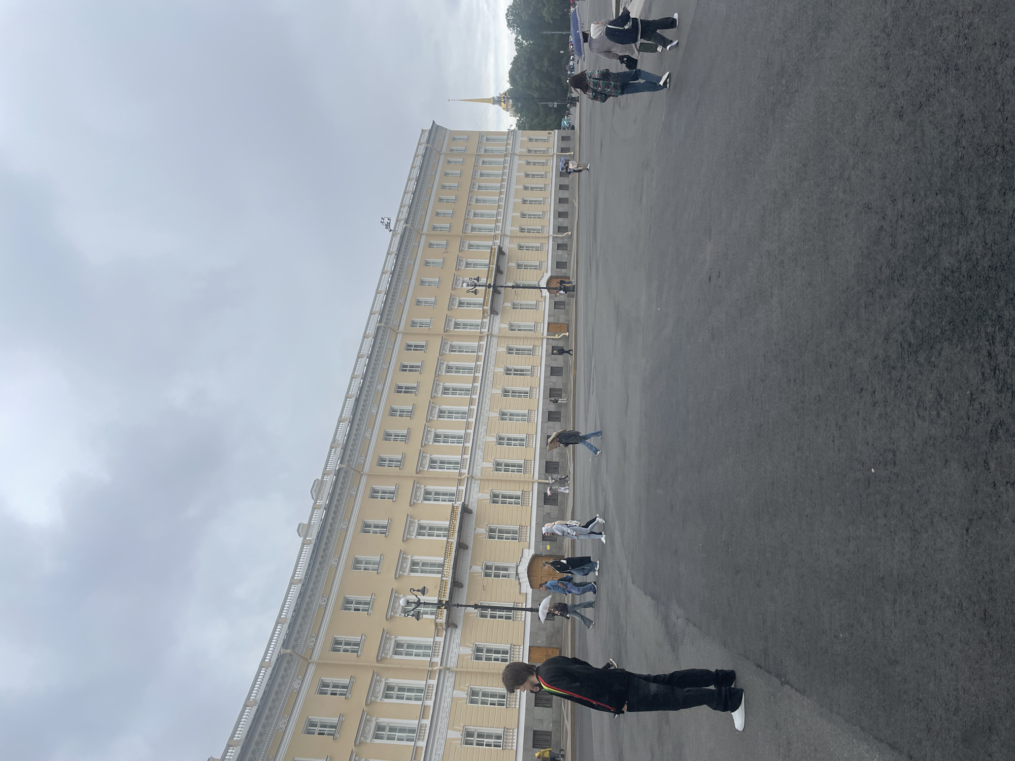 The image depicts a wide, open paved area in front of a large, pale yellow, neoclassical building. The building has multiple floors and numerous windows, with decorative white detailing along the roofline and between floors. A streetlamp with an ornate design stands in the foreground. Several people are walking in the distance, some holding umbrellas, suggesting overcast or rainy weather. On the far right, a tall, slender golden spire topped with a cross pierces the cloudy sky, a distinctive landmark of Saint Petersburg, Russia. The foreground shows a small section of the pavement and the lower body of a person standing facing away from the camera, wearing black pants and white sneakers. The overall scene suggests a public square in a historic urban setting.
