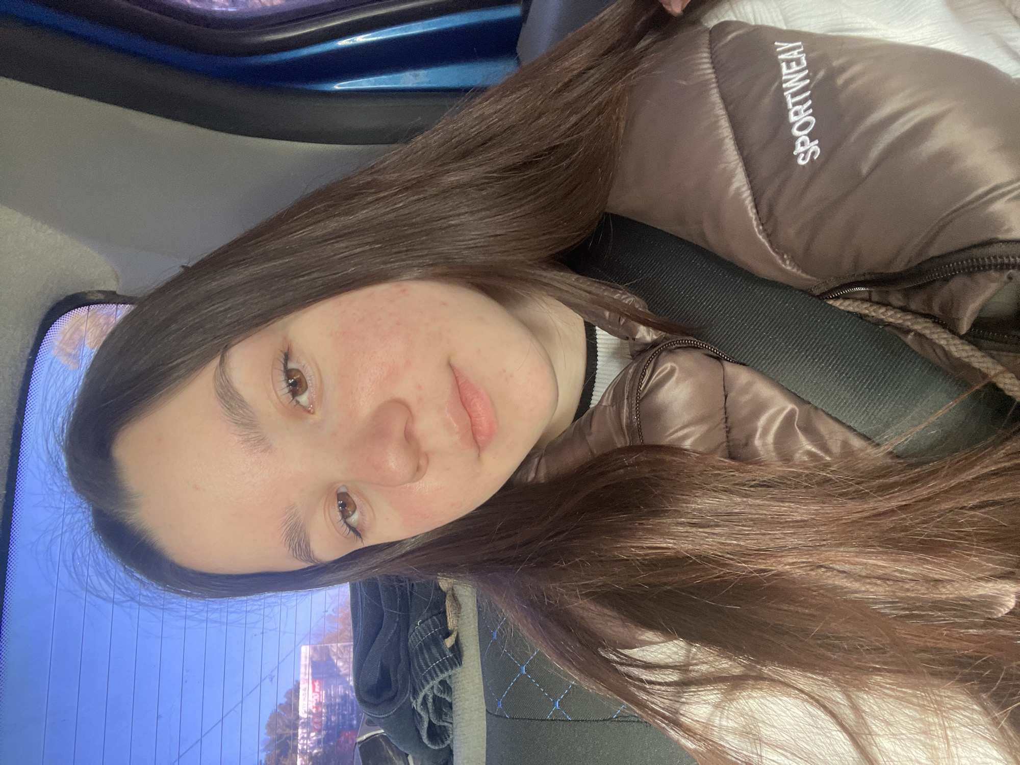 A young woman with long brown hair is sitting in a car. She is wearing a brown puffer jacket with the word "SPORTWEAY" visible on the upper right side. She has a seatbelt on. Her face is visible, and she has some redness on her cheeks. The car appears to be moving, based on the blurred scene visible through the rear window. The location is Chaikovsky, Russia, as indicated by the view through the rear window which includes street elements and buildings characteristic of a Russian city. The car's interior includes a seat with blue stitching, and the exterior is blue.