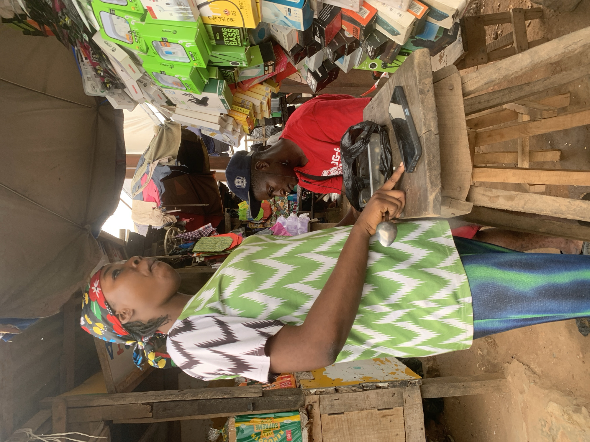 A medium shot captures a busy outdoor market scene in Shagamu, Nigeria, focusing on two individuals engaged in a transaction. On the left, a woman wearing a vibrantly patterned headwrap and a green and white zig-zag patterned top is seen from the chest down, holding a metallic object in her right hand. Her left hand rests on a wooden surface. To her right, a man in a red t-shirt and a dark baseball cap sits behind a wooden counter laden with electronics accessories. He is looking down, seemingly interacting with the items on the counter. The background is filled with shelves stacked with various boxed electronic accessories, suggesting a mobile phone or accessories stall. The ground appears to be dusty earth. The lighting suggests it is daytime, likely under the cover of tarps or makeshift roofing.