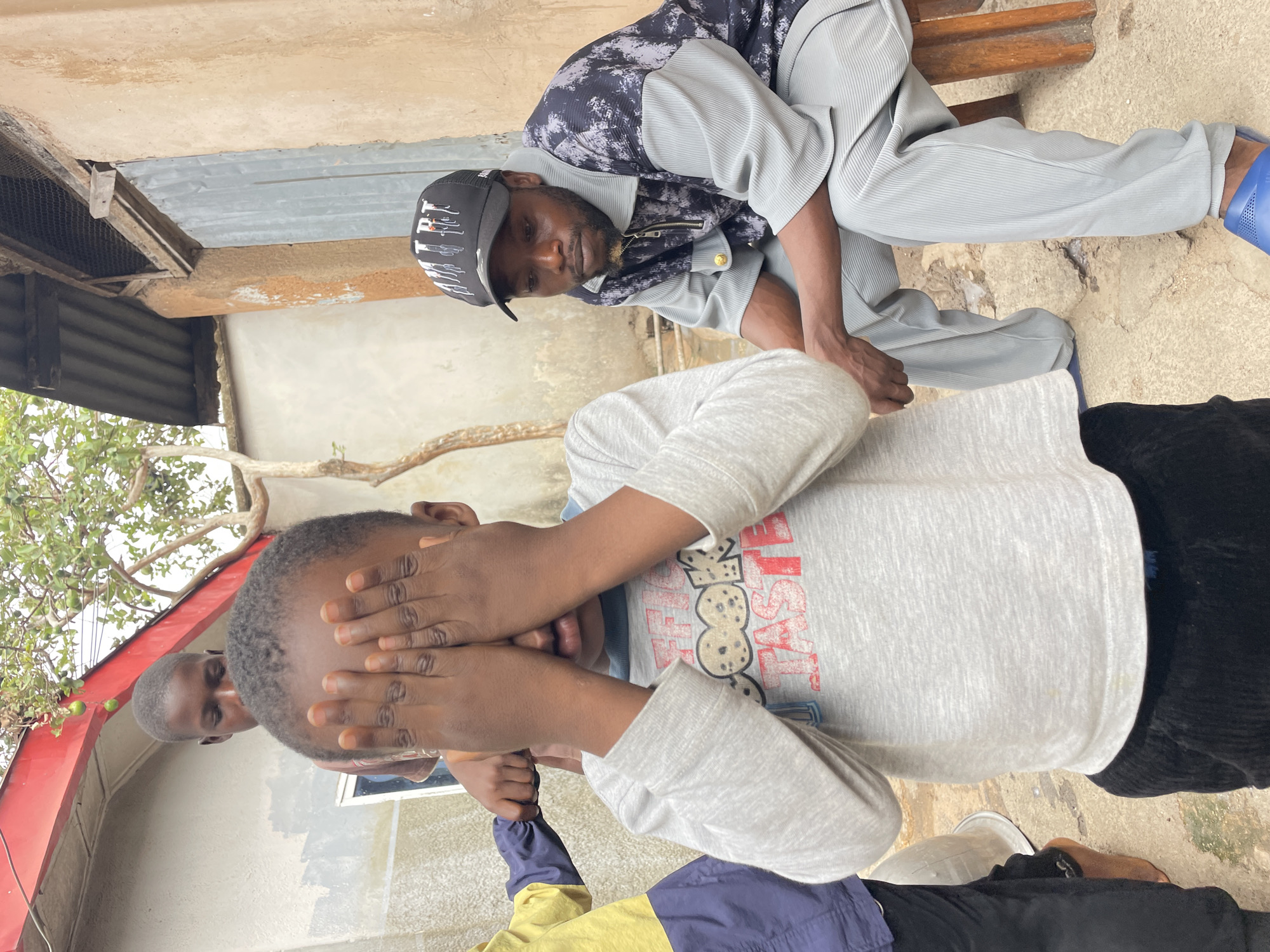 This image captures an outdoor scene in a residential area of Jos, Nigeria, featuring four individuals. In the central foreground, a young, dark-skinned child wears a light gray long-sleeved shirt with "COOK TASTE" lettering and a graphic, paired with black corduroy pants. Both of the child's hands are placed over their face. To the left, a second child, partially visible, wears a yellow and dark blue jacket with black pants; their right hand is in contact with the central child's left arm, and their left foot is bare. A third child is visible in the background, behind the central child, looking downwards. On the right side of the frame, an adult male with facial hair is seated on a low wooden stool. He wears a black baseball cap with white text and graphics, a short-sleeved, collared shirt that is dark patterned on top and light gray ribbed on the bottom, light gray ribbed pants, and blue plastic slide sandals.
The environment includes two visible structures: a light gray building with a red roof edge and a window on the left, and a textured light brown building with a corrugated metal door and wire mesh above it on the right. Green foliage, including a tree bearing small green fruits, is present near the left building. The ground consists of uneven concrete and packed earth, with a white basin partially visible on the left. The lighting is diffused, indicating an overcast sky or shaded conditions.