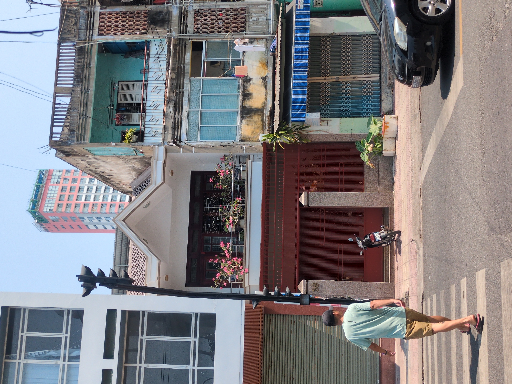 The image depicts a street scene in Nha Trang, Vietnam. A man wearing a light green t-shirt, khaki shorts, a dark cap, and flip-flops is walking across a crosswalk. A parked motorbike is adjacent to a building with a red corrugated metal gate. Behind the gate, a two-story building with a white façade, red tiled roof, and window boxes with pink flowers is visible. Adjacent to this building is a faded green and blue multi-story building. A black car is partially visible parked along the sidewalk. A tall pink and red building is in the background. The day appears bright and sunny.