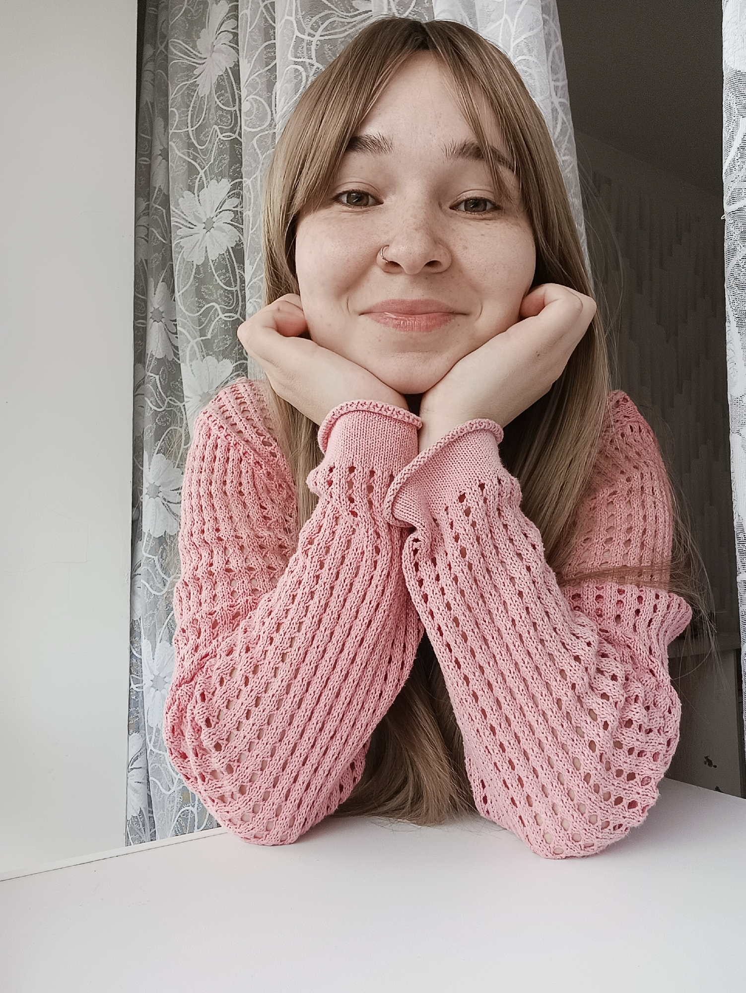 A young woman with light brown hair and freckles is posed indoors, resting her chin on her hands. She is wearing a pink knitted sweater with decorative sleeves. Behind her is a window with a sheer, floral-patterned curtain. The surface in front of her is white. The location of the image is unknown; Izhevsk, Russia, cannot be confirmed.
