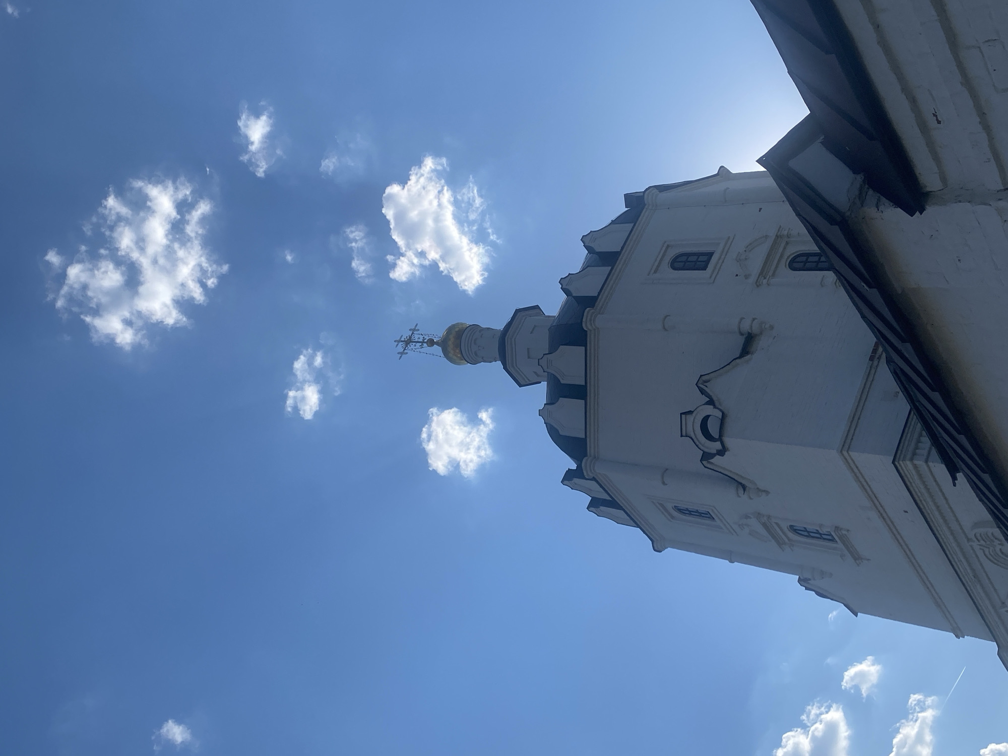 The image shows the architectural details of a building in Sviyazhsk, Russia, featuring a white tower with a golden dome topped by a cross. The tower is set against a bright blue sky with scattered white clouds. A portion of the building's roofline is visible in the upper right corner of the image.