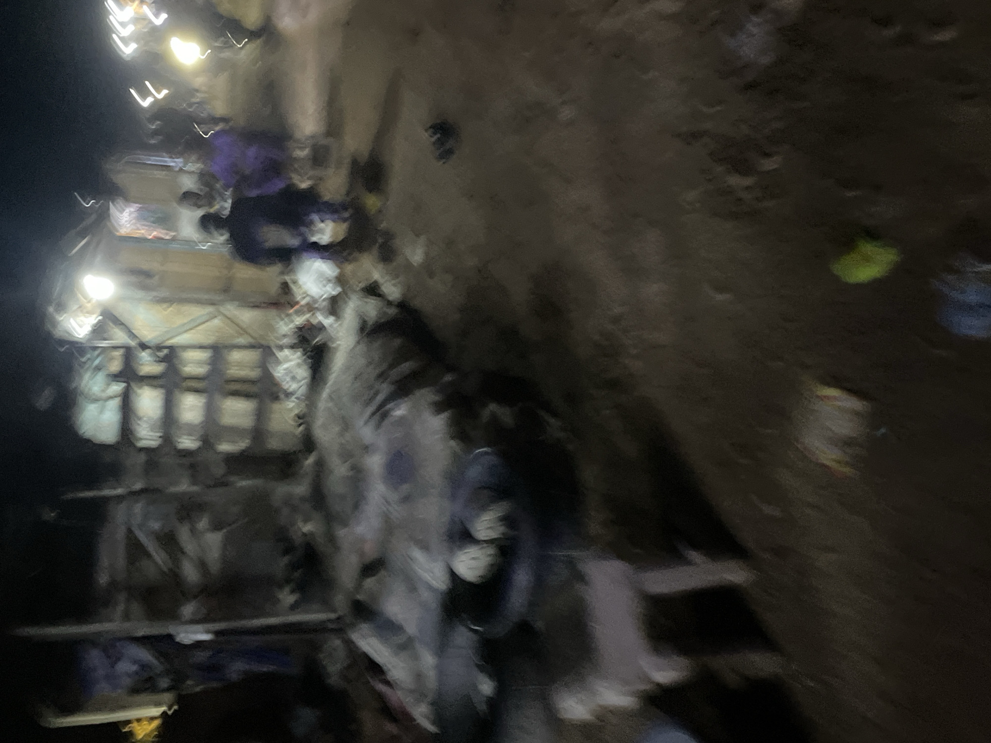 The image depicts an outdoor scene at night, likely in a low-income urban area of Lokoja, Nigeria, with visible makeshift structures and scattered debris. In the foreground, a dark, possibly circular object, possibly a tire, is visible with two lighter-colored round objects resting on it, suggesting food items or containers. To the left, a portion of a light-colored, stool-like object is present. Further back, several figures are blurred due to low light and motion. One figure in the center appears to be wearing a dark garment, possibly a jacket, and another to their right is in a purple garment. To the left of the central figures, another individual in a dark garment is seen. The background features various structures, including what looks like corrugated metal sheeting and possibly tarpaulin. Illumination comes from bright, diffused light sources, likely lamps, casting stark highlights and deep shadows. The ground appears to be dirt or packed earth, littered with unseen debris. The overall impression is of a bustling, dimly lit environment, potentially a market or residential area at night.