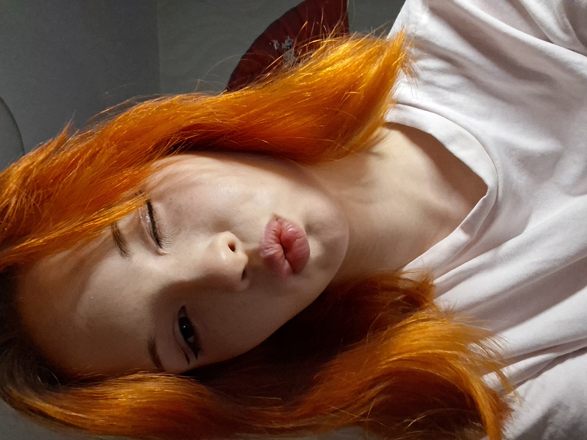 The media file presents a close-up, eye-level portrait of a person with striking, vivid orange hair, lying down and looking towards the viewer. The person's head is tilted, and their body is angled diagonally across the frame. Their lips are pursed in a "kissy" face or pout, and their right eye is mostly closed, while the left eye is partially open, showing a dark pupil and some eyeliner.
The person has fair skin, with soft highlights and shadows on the face. They are wearing a light-colored, possibly white or very light pink, plain t-shirt or top with visible folds in the fabric. A dark red, fan-like object is partially visible behind the person's head, amidst the bright orange hair, contrasting with the muted, dark grey-green background.
The setting appears to be indoors, likely in Chaikovsky, Russia, with soft, ambient lighting. No specific time of day or weather conditions are discernible from the image, nor is there any visible text. The overall mood conveyed by the pose and expression is relaxed and playful.
