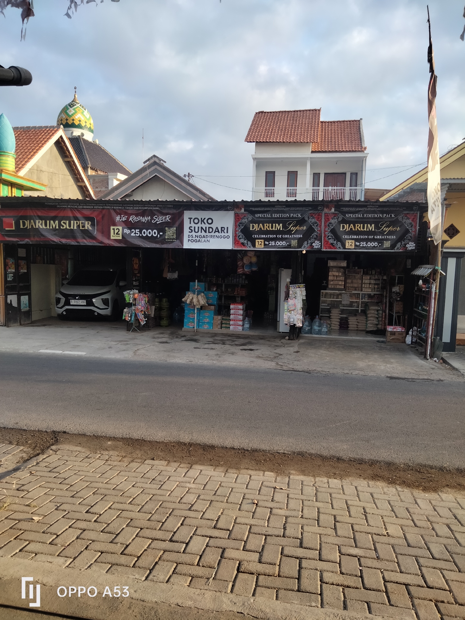 The image shows a storefront in Trenggalek, Indonesia, identified by the presence of a white car with license plate B 1842 JQC parked inside and a sign in the foreground that reads "OPPO A53". The storefront itself is advertising "Djarum Super" cigarettes with a special edition pack for Rp 25,000. There are also displays of various goods for sale inside the store, including what appear to be bags of rice, bottled water, and other packaged items. The building has a traditional Indonesian architectural style with a tiled roof and an outdoor awning. A mosque with a green and yellow dome is visible in the background, as is a white multi-story building with a red tiled roof. The street in front of the store is paved with interlocking bricks, and there is a sidewalk separating the street from the store's entrance. The sky is partly cloudy.