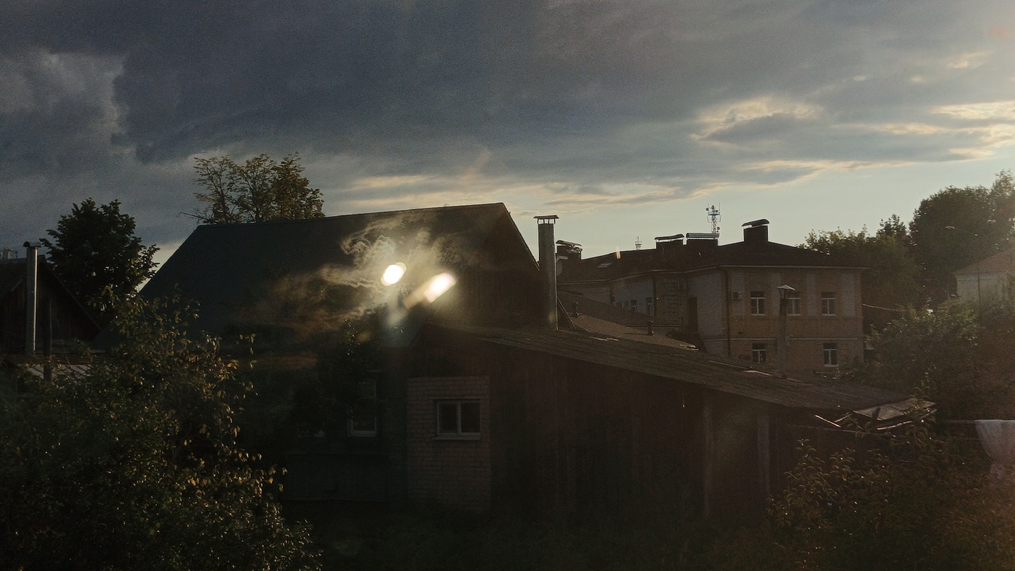 This image depicts a residential area in Semyonov, Russia, likely during dusk or dawn, given the dramatic interplay of light and shadow. The foreground is dominated by the dark silhouette of a wooden structure with a sloping roof. Two bright, diffused light sources, possibly reflections or a light source obscured by atmospheric conditions, shine from the roofline of this structure, creating a hazy, almost ethereal effect. A chimney rises from this structure, a metallic pipe.
Behind the foreground structure, a larger, darker roof of another building is visible, also with a chimney. Further in the background, a light-colored apartment building with multiple windows and several chimneys stands under a turbulent, dark gray sky. The sky is illuminated by horizontal bands of lighter clouds and a soft glow of sunlight near the horizon. Trees and foliage are present in the midground and foreground, adding to the natural, perhaps rural or suburban, feel of the scene. The overall impression is one of a quiet, somewhat moody, possibly post-industrial or older residential neighborhood.