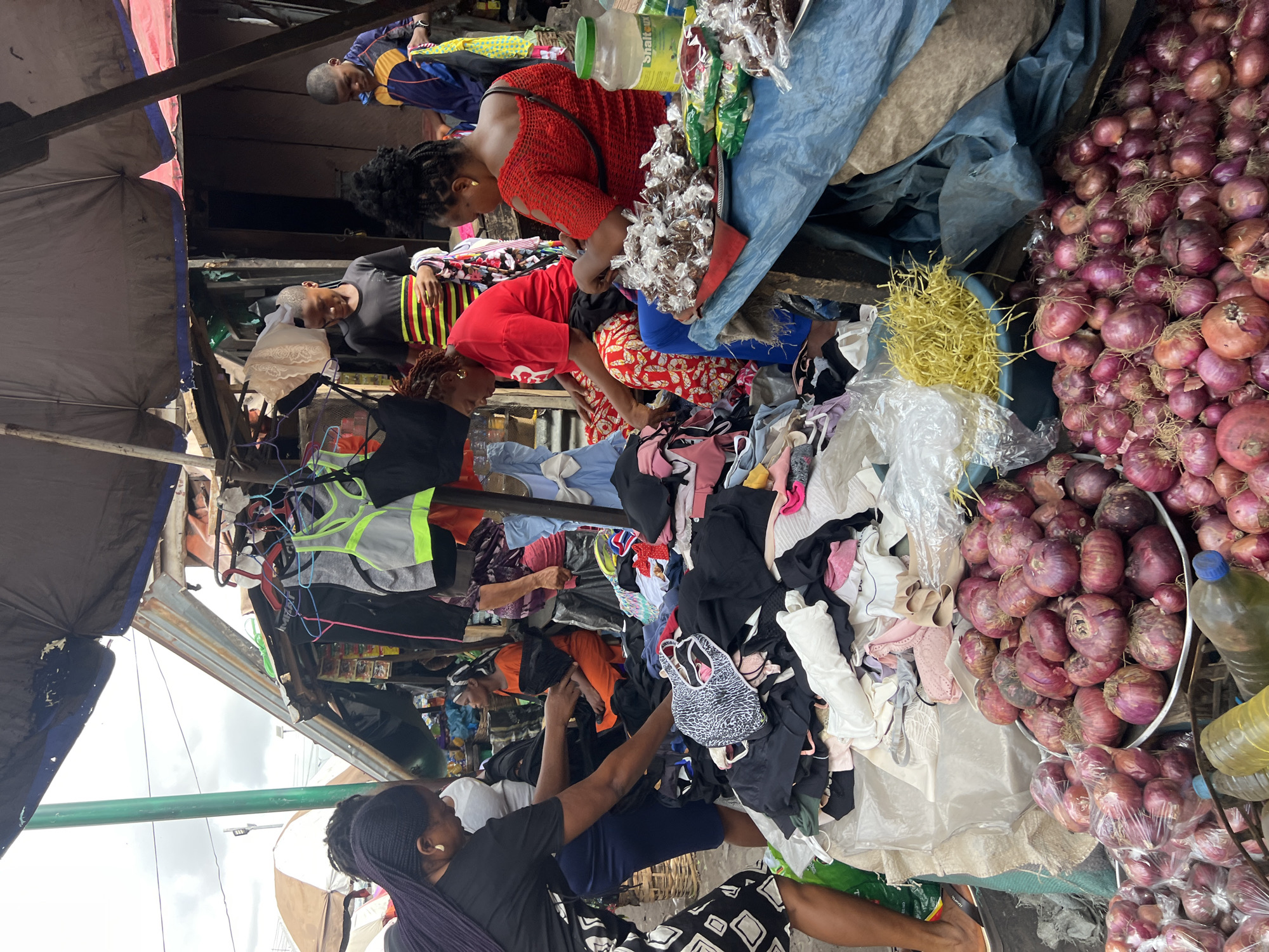 The image captures a vibrant and busy open-air market scene in Nkpor, Nigeria. The photograph is presented rotated 90 degrees counter-clockwise.
The market is bustling with several individuals, primarily women, engaged in shopping and selling activities. In the upper central portion of the image (when correctly oriented), a woman with dark braids and a black top is seen examining goods. To her right, a man in a blue and yellow jacket is visible in the background. Further down in the central area, a woman in a bright red, textured top leans over, intently inspecting merchandise, with a green-capped plastic container nearby bearing the partially legible text "Shaltea." Below her, another woman in a red t-shirt is bent over a large pile of clothes, and another woman sorts through a heap of garments.
The market stalls showcase a variety of goods. A prominent feature on the right side of the frame (when correctly oriented) is a substantial mound of red onions, some of which are pre-packaged in clear plastic bags, indicating a produce stall. Nearby, various plastic bottles, one with yellow liquid and another with a blue cap, are visible. Large quantities of clothing items, including bras, sports tops, and other garments, are displayed on tables or piled for customers to browse. Small, clear plastic bags filled with what appear to be snacks or sweets hang near one of the stalls. A blue bin contains yellow, straw-like material.
The market environment features makeshift structures, with tarpaulins, umbrellas, and corrugated metal sheets forming coverings over the stalls to provide shade. An overcast sky is visible in some sections, indicating it is daytime. The atmosphere is one of focused commerce, with individuals absorbed in the process of buying and selling, characteristic of a dynamic local market.