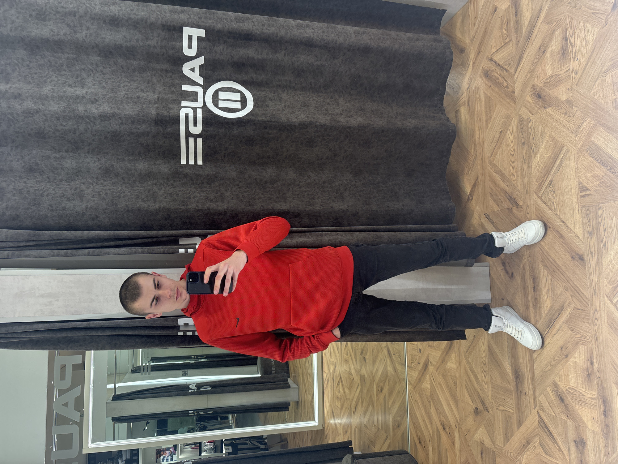 A young man with a shaved head stands in what appears to be a clothing store dressing room. He is wearing a red hooded sweatshirt, black jeans, and white sneakers. He is holding a black smartphone and taking a selfie. The dressing room has a mirror on one side, reflecting the interior of the store. There are gray curtains with the word "Pause" and a circle with two vertical lines inside, visible behind him. The floor is patterned with wooden planks. The location is Plovdiv, Bulgaria.