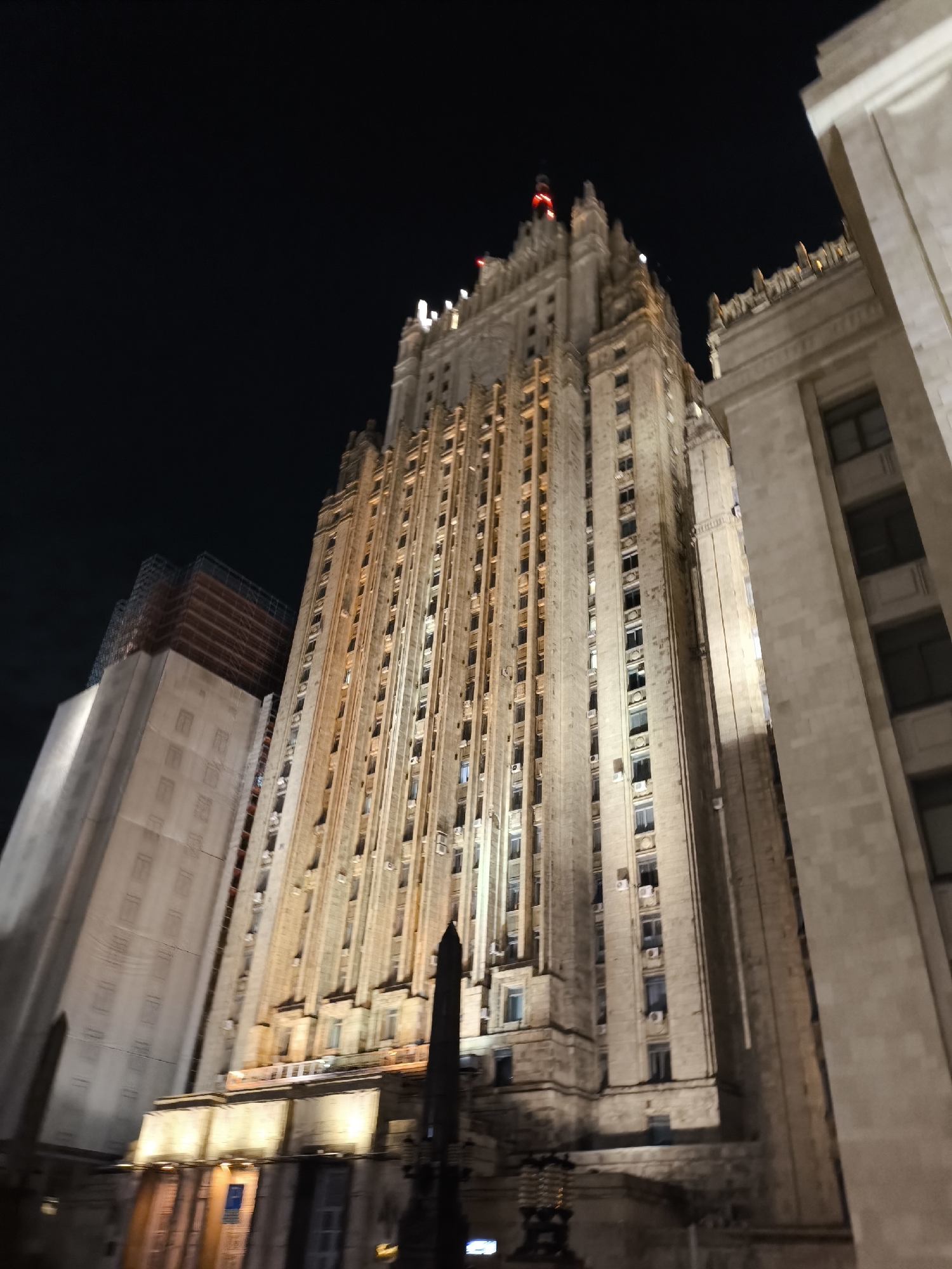 The image captures a towering, ornate building illuminated against a dark, clear night sky, viewed from a low-angle perspective. The main subject is a monumental edifice exhibiting characteristic Stalinist Empire architecture, with numerous vertical elements, stacked tiers, and a prominent spire-like top featuring red lights. Warm, yellowish artificial light highlights the intricate details of its stone facade, which includes many windows and various decorative features.
To the right of the main building, a section of another large, similarly constructed building with a lighter facade is visible, also illuminated. On the left, a third structure, appearing less detailed and partially covered by scaffolding on its upper levels, suggests ongoing construction or renovation.
In the foreground, a dark, pointed, possibly metallic object, likely a monument or an architectural feature, rises partially obscuring the lower part of the main building. The absence of daylight and the artificial illumination confirm the nighttime setting. No individuals or specific activities are depicted. The architectural style and scale strongly suggest the location to be Moscow, Russia, aligning with the "Seven Sisters" skyscrapers, such as the Ministry of Foreign Affairs building. No discernible text is visible on any of the structures.