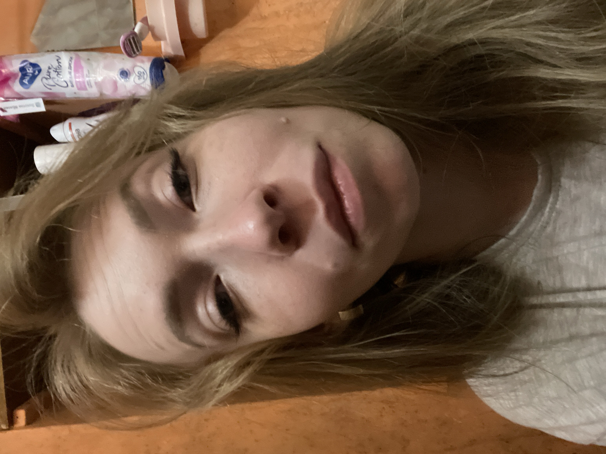 A close-up, angled shot shows a woman with light blonde hair and fair skin. Her face is the primary focus, with her eyes appearing dark with makeup and her lips slightly parted. A small mole is visible on her forehead.
In the background, various personal care items are scattered, including a bottle labeled "Aura Pure Cotton".
It is not possible to determine the location of this image as Perm, Russia, or any specific location from the provided image alone.
