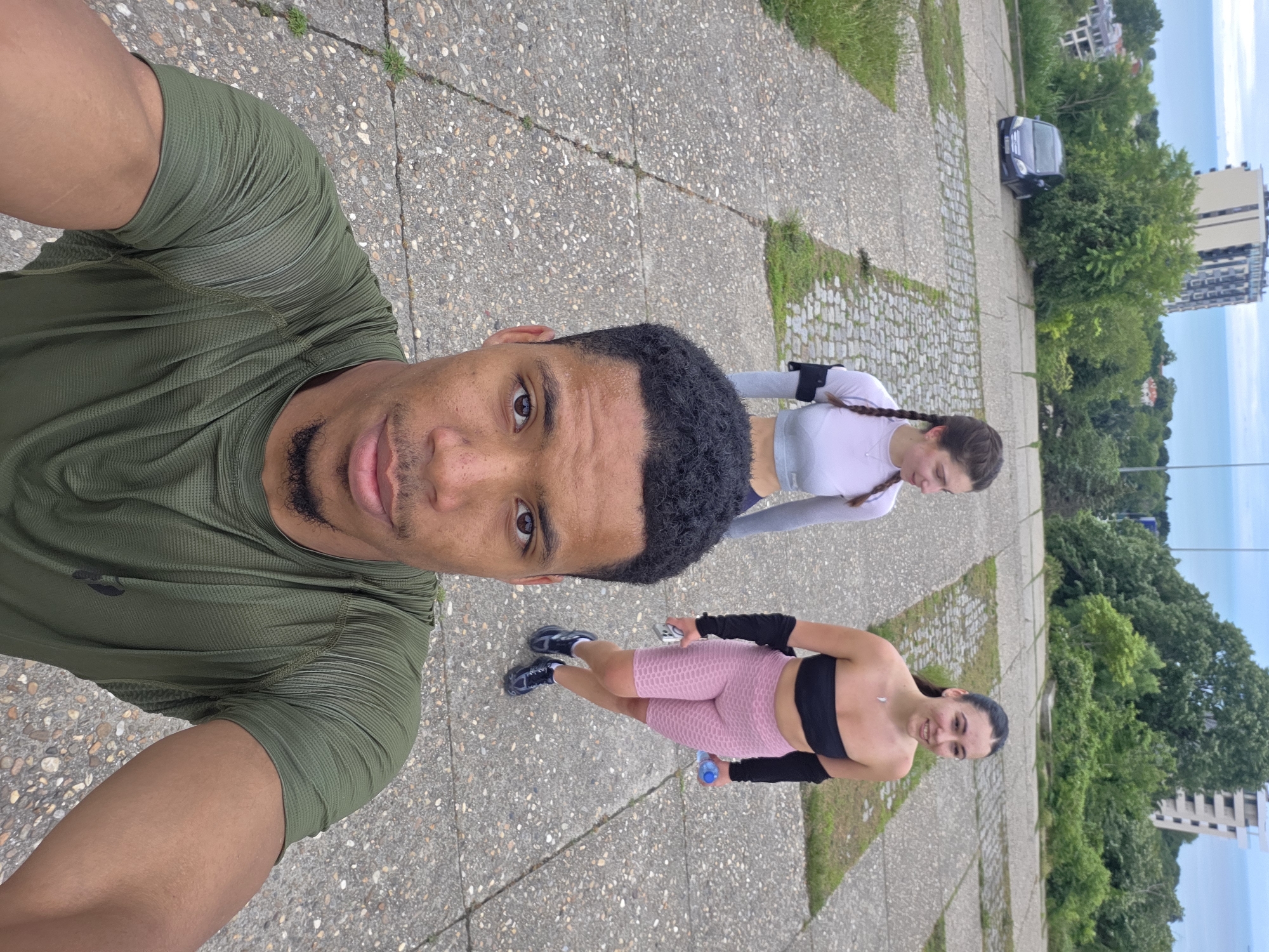 In Varna, Bulgaria, three individuals are walking up a concrete pathway on a cloudy day. In the foreground, a Black male is taking a selfie, wearing a green t-shirt. Behind him, two white females are walking away from the camera. The female on the left is wearing a light purple crop top and the one on the right is wearing a black bandeau top and pink shorts. In the background, there are trees, buildings, and the Black Sea.