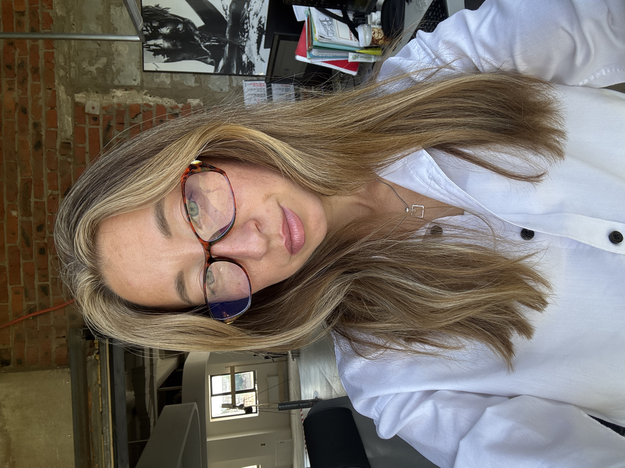 This is a selfie of a woman wearing a white collared shirt and glasses, taken indoors. She has long, blonde hair and is looking at the camera. The background appears to be an office space with exposed brick walls and industrial-style lighting fixtures. There are windows in the background showing an outdoor scene. A desk with a computer and various office supplies is visible to the right. The image is not set in the city of Chaikovsky, Russia, as it does not contain any specific identifiable elements of that location.