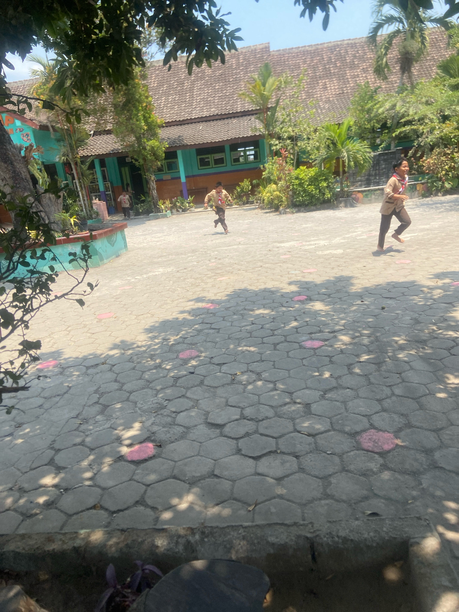 The image shows an outdoor scene in front of a school building in Sleman, Indonesia. Two young boys are running across a paved courtyard. They are both wearing brown uniforms with red and white scarves. The school building has a tiled roof and teal-colored walls with green window frames. There are trees and lush green plants surrounding the building and courtyard. The courtyard is paved with hexagonal concrete blocks, and there are several pink circular markers on the ground. The sun is shining brightly, casting shadows from the trees onto the ground.