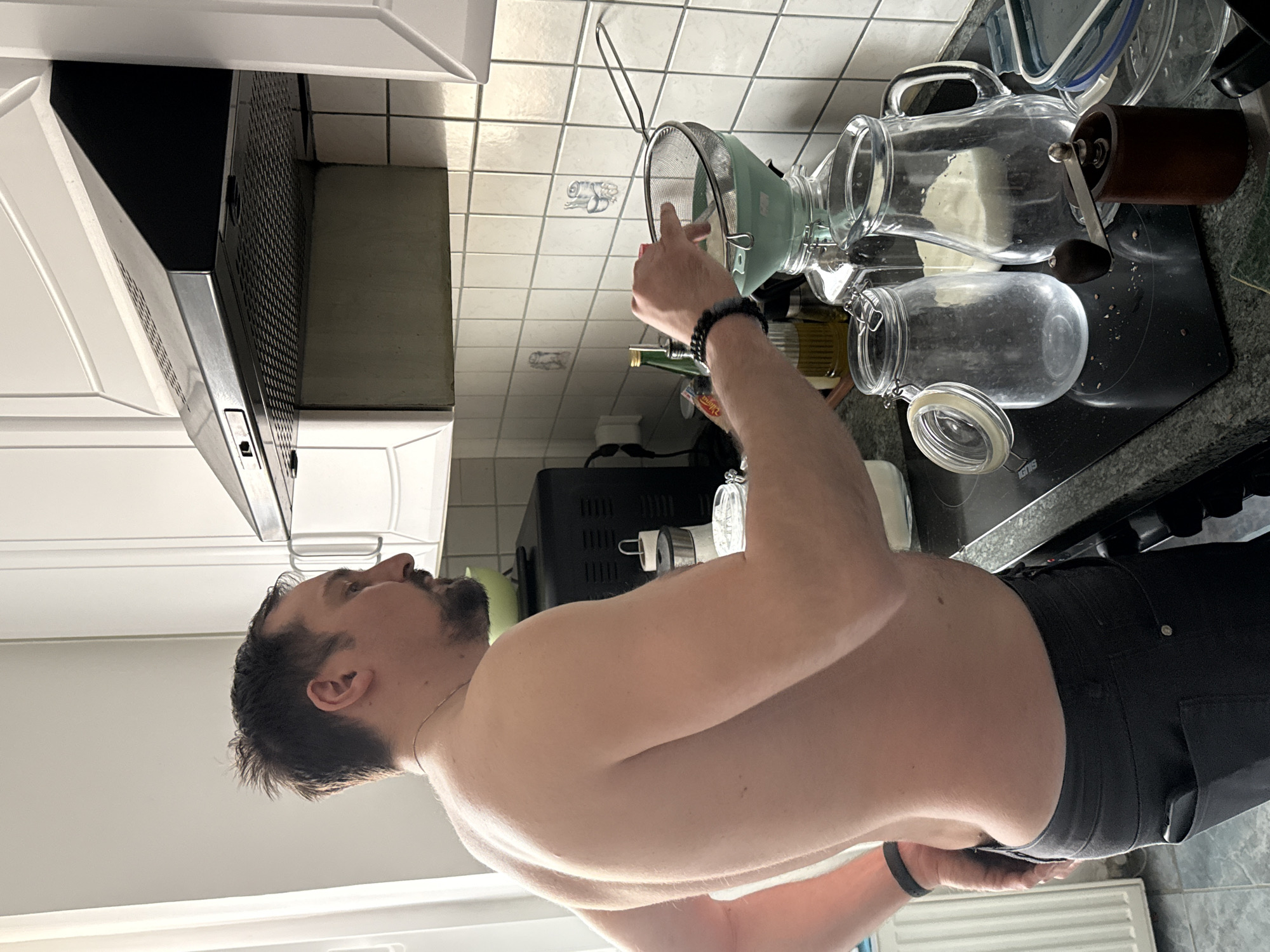 This medium shot captures a man in a domestic kitchen, engaged in a culinary task. The man, shirtless with dark hair and a beard, is visible from the chest up, focused on an activity at the countertop. He is wearing dark trousers and a black wristband on his left arm.
The setting is a well-lit kitchen featuring white upper cabinets, a black range hood, a white tiled backsplash, and a dark granite-like countertop. A black induction cooktop, with the brand "IGNIS" faintly visible, is situated on the counter.
The man's right hand holds a light green, bowl-shaped strainer over an open clear glass jar, suggesting he is pouring or straining a liquid or fine solid. Nearby on the counter are several other glass containers, including a large pitcher and another swing-top jar. A manual coffee grinder with a wooden handle is positioned next to the pitcher. An olive oil bottle and a black kitchen appliance are also present. The white tiled backsplash exhibits subtle, light grey outlines of kitchen-related drawings, such as a rolling pin and cutlery. A white radiator is visible at the bottom of the frame.
The man's expression is neutral, conveying focus on the task. The lighting suggests a daytime setting. No specific visual cues within the image confirm the provided location context of Hainburg an der Donau, Austria.