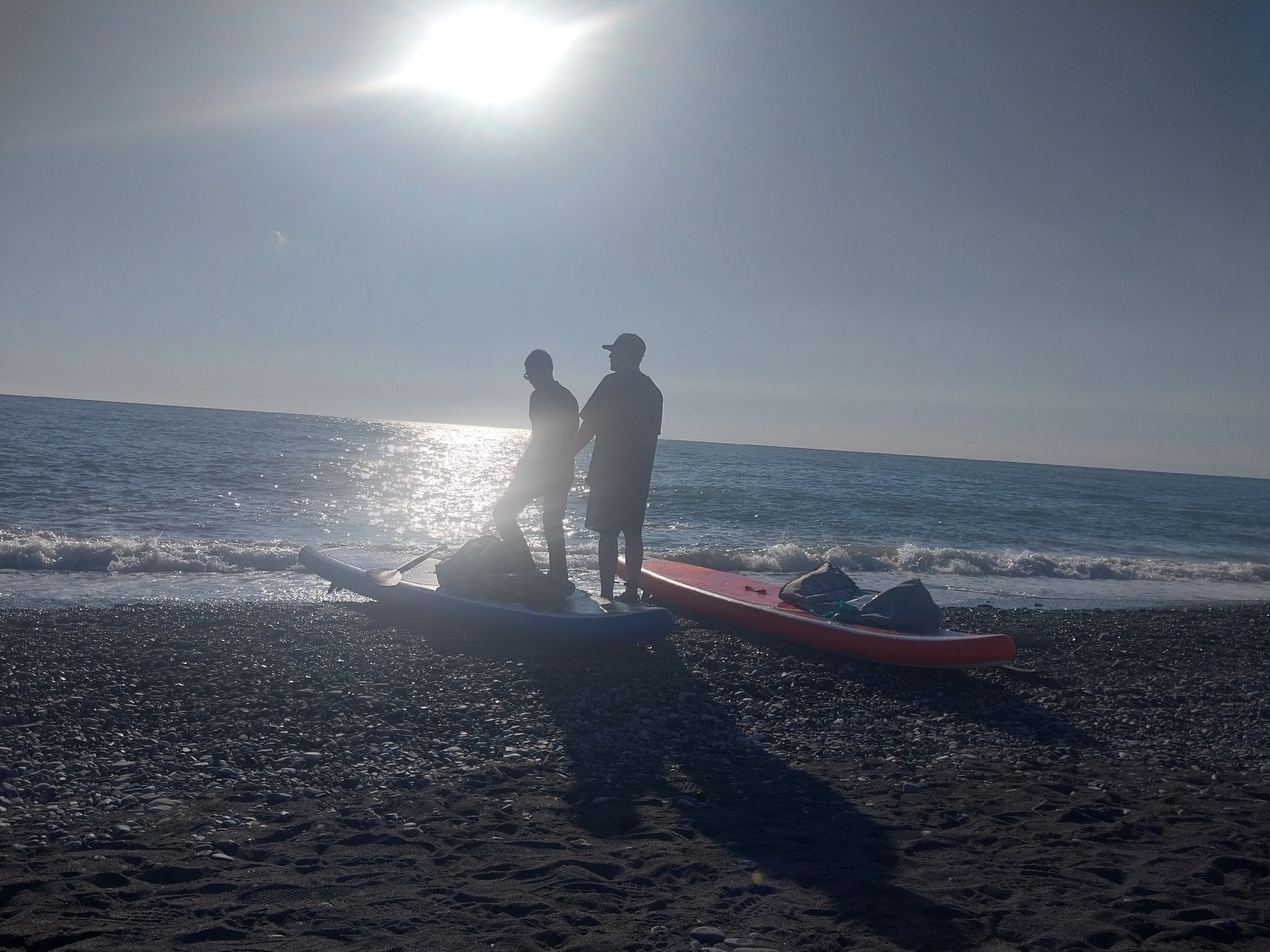 This media file captures a sun-drenched coastal scene, likely in Sochi, Russia, featuring two individuals and their paddleboards on a pebble beach.
The dominant subjects are two silhouetted figures, appearing to be men, standing near the water's edge. The bright, low sun behind them creates a strong backlight, making their features indistinct and casting long shadows across the dark shingle and sand. One person is positioned next to a blue paddleboard, which has a paddle and some gear on it. The other person stands closer to a red or orange paddleboard, which is further up on the beach and appears to have bags or equipment secured to it. The individuals seem to be preparing for or concluding a paddleboarding session.
The setting is a tranquil beach with small, dark pebbles and patches of sand. Gentle waves are breaking on the shore, reflecting the intense sunlight as sparkling highlights. Beyond the beach, the vast expanse of the sea stretches to the horizon under a clear, bright sky. The sun is prominently visible in the upper center of the frame, causing significant lens flare.
The time of day is either early morning or late afternoon, given the low angle and intensity of the sun. The weather appears to be clear and sunny. No visible text is present in the image.