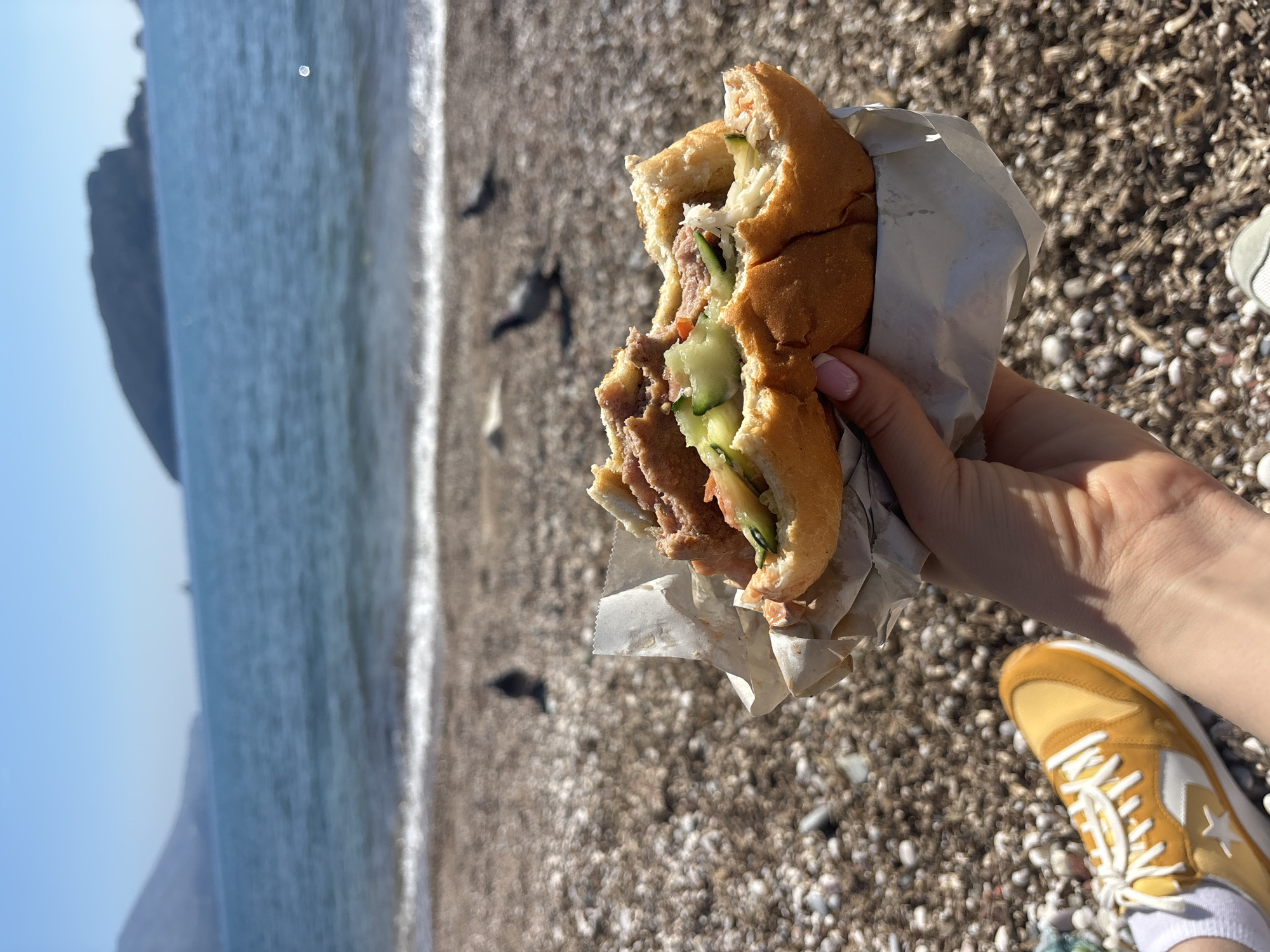A person holds a half-eaten sandwich in a paper wrapper in front of a pebbled beach, with a body of water and a cliff in the background. The sandwich contains meat and cucumber slices. The person's lower arm, hand, and a yellow and white sneaker are visible. The location appears to be the beach in Budva, Montenegro.