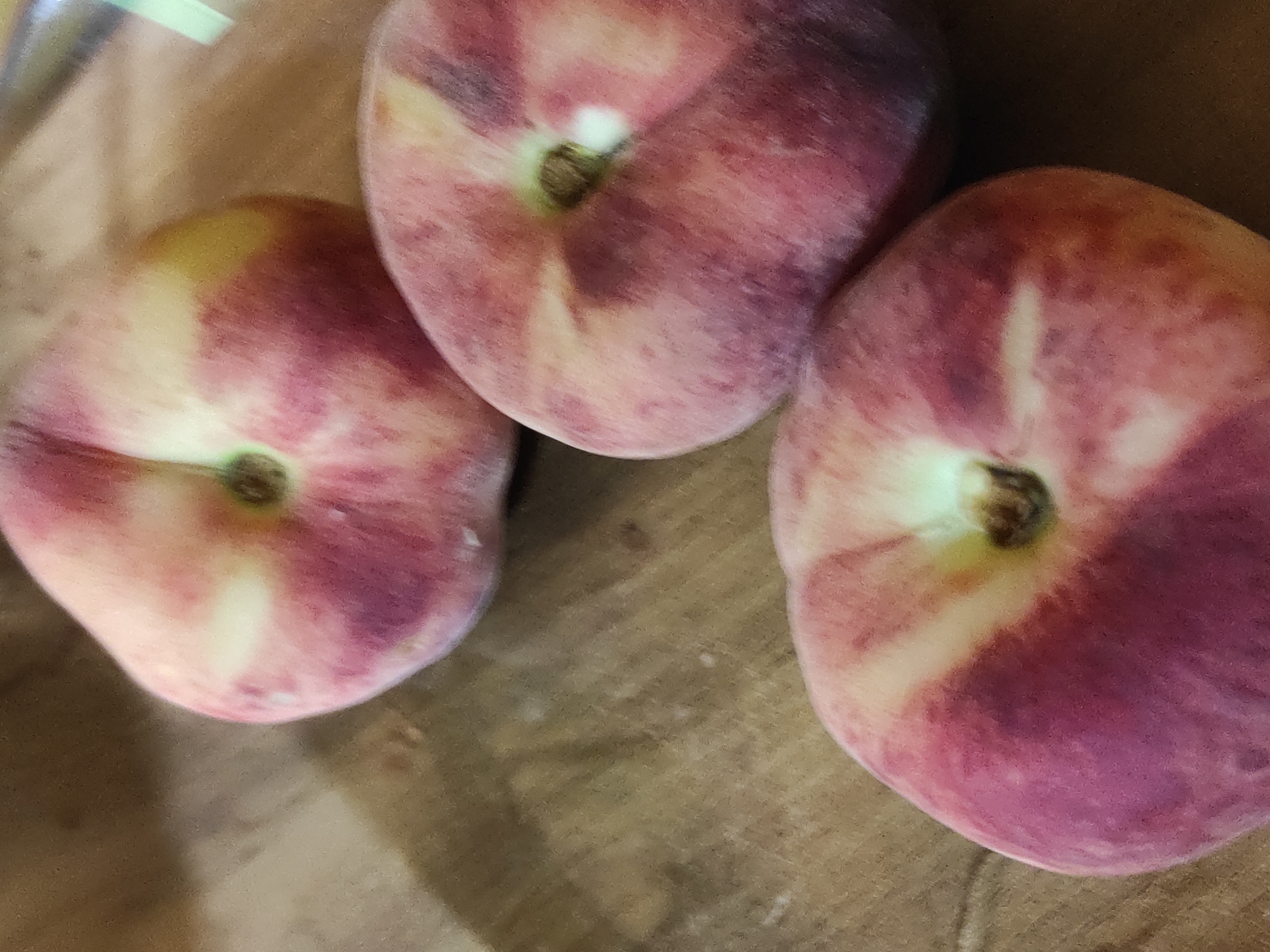A close-up shot showcases three flat peaches resting on a textured, wooden surface. The peaches are round with a distinct indentation, characteristic of flat varieties. Their skin displays a gradient of colors, ranging from a soft yellow near the center to a deep pink and reddish-purple on the outer edges. The surface beneath them has a grainy, wood-like texture, appearing to be a table or countertop. The lighting is warm, casting soft shadows and highlighting the fuzzy texture of the peaches. The overall impression is one of fresh fruit, arranged simply on a natural surface. The image appears to have been taken indoors, with no discernible elements of an outdoor environment or specific urban features.