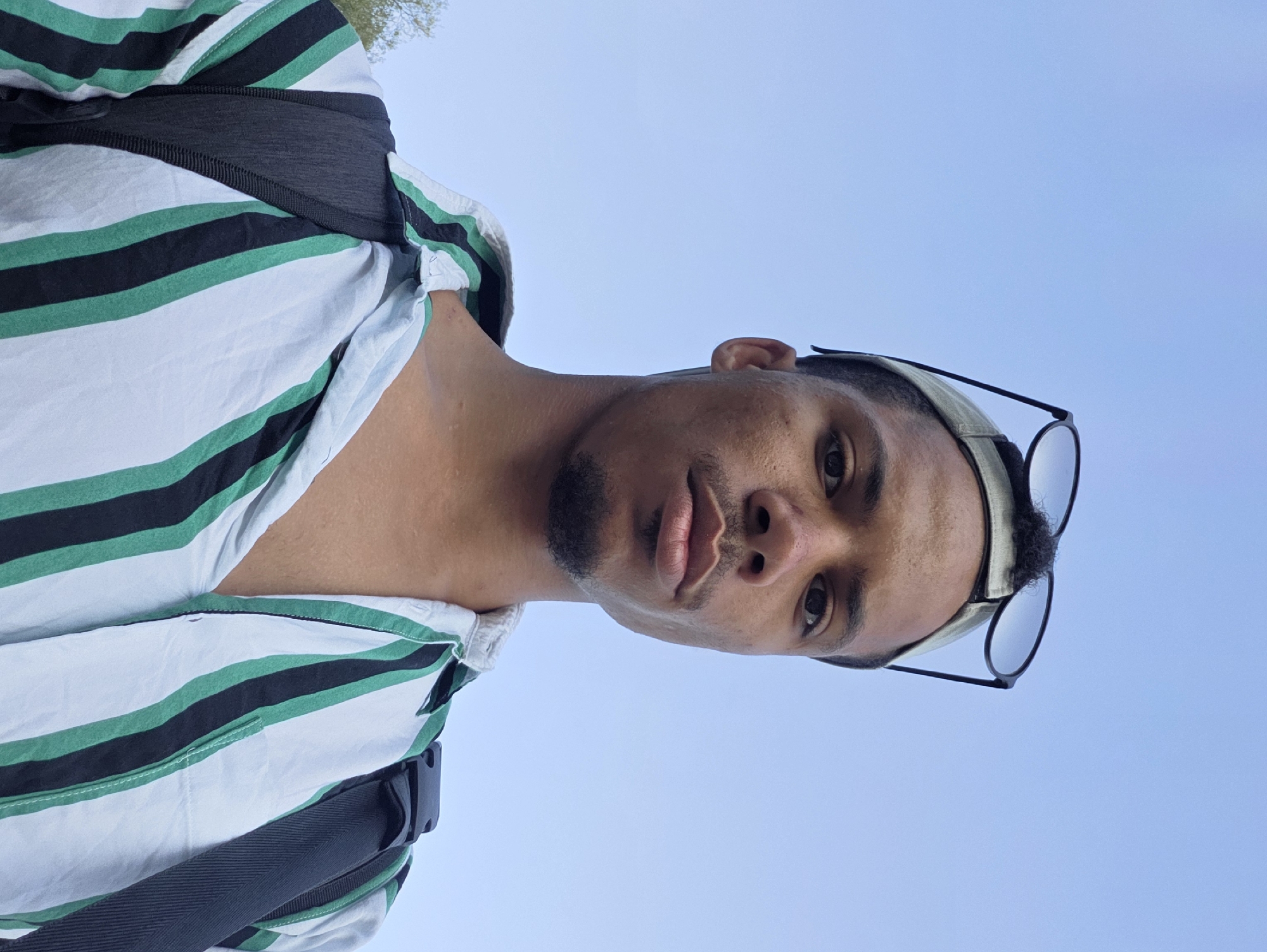 A medium shot selfie of a young Black man outdoors, looking directly at the camera. He is wearing a striped collared shirt with green, black, and white stripes. A backpack is visible with black straps. A baseball cap is on his head, with sunglasses resting on top of the cap. The man has a short beard and a goatee. The background is a clear blue sky with some faint greenery visible on the left side, suggesting an outdoor park or natural setting. The image was taken during the day. The location is not specified.