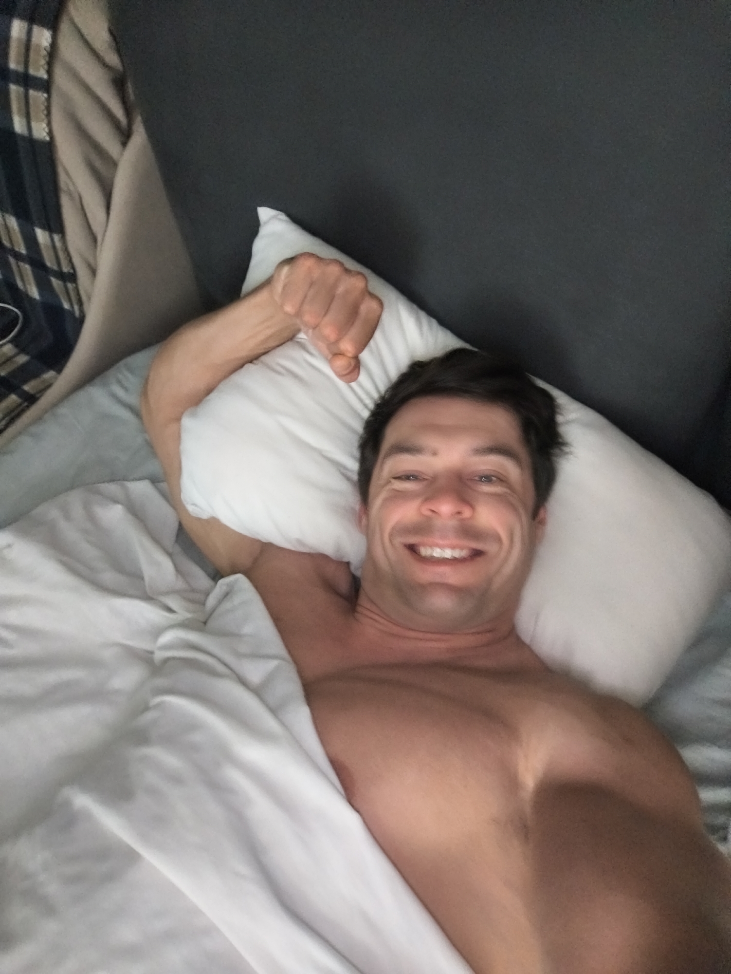 A shirtless man is lying in bed with a white sheet partially covering his torso. He is smiling and looking at the camera. His right arm is raised with a clenched fist resting on a white pillow. The bed has a gray headboard. There is a plaid blanket visible on the left side of the image, near a light-colored cushion. The location is not identifiable as Moscow, Russia.