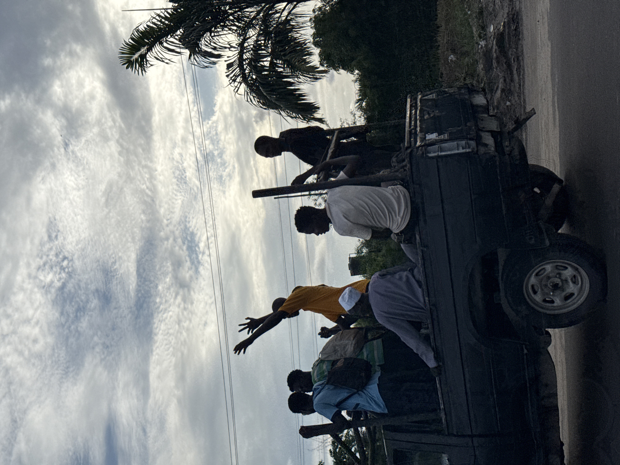 Several people are riding in the back of a dark-colored pickup truck. The truck is on a road in Lekki, Nigeria. The sky is cloudy. A palm tree is visible above the truck. The people in the truck are silhouetted against the sky. One person is reaching out with an outstretched hand.