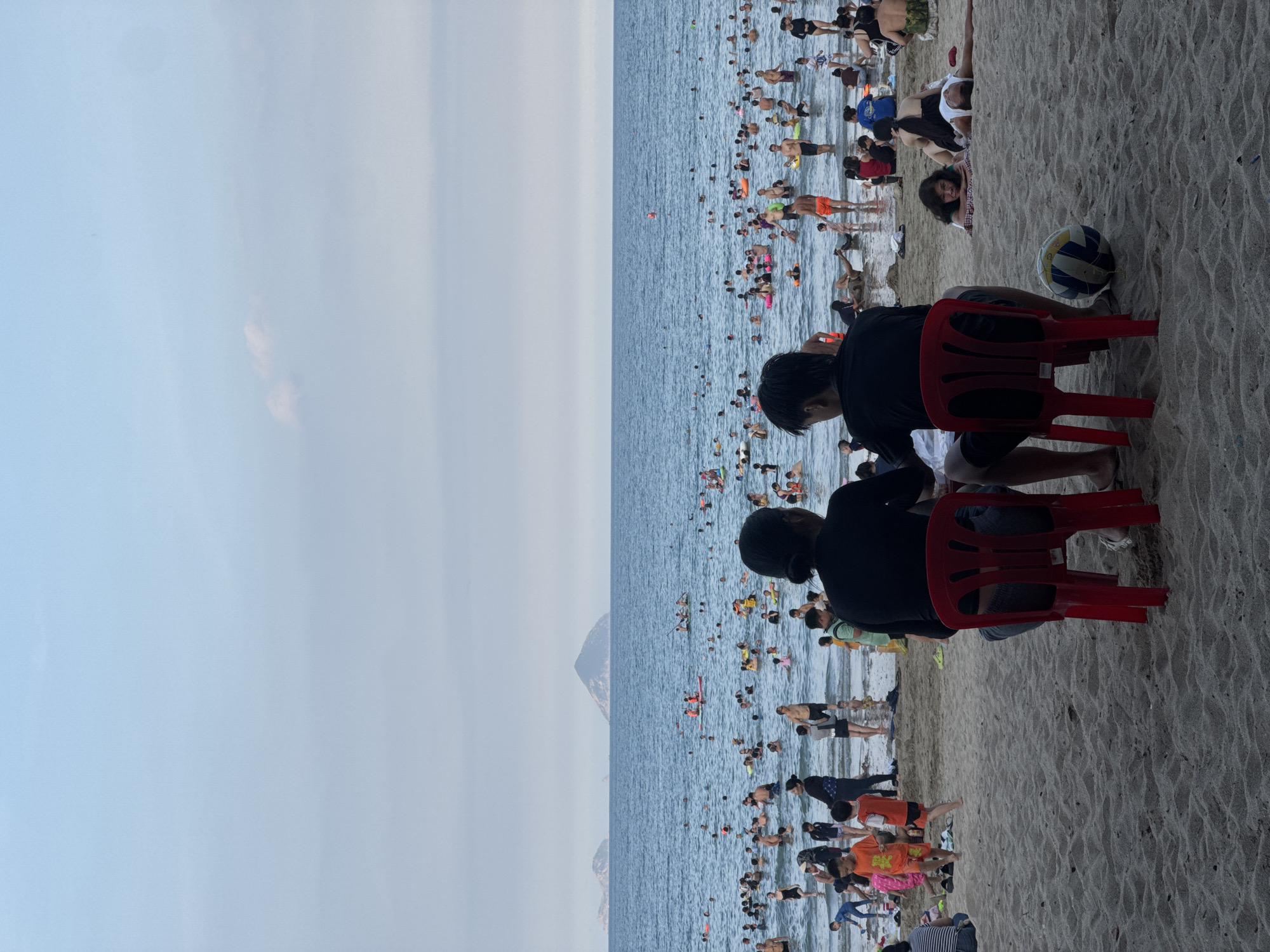 The image depicts a crowded beach scene in Bac Nha Trang Ward, Vietnam, during daylight hours. The ocean is a deep blue, with numerous people swimming and wading in the water. The water's edge is bustling with activity, with individuals of all ages enjoying the beach. In the foreground, two figures are seated on red plastic chairs facing the ocean, suggesting they are observing the activity or resting. A volleyball rests on the sand near their feet. The beach itself is sandy, with gently rolling dunes. In the background, several islands rise from the ocean, adding to the scenic landscape. The sky is a pale blue with some faint clouds. The overall impression is one of a lively and popular tourist destination.