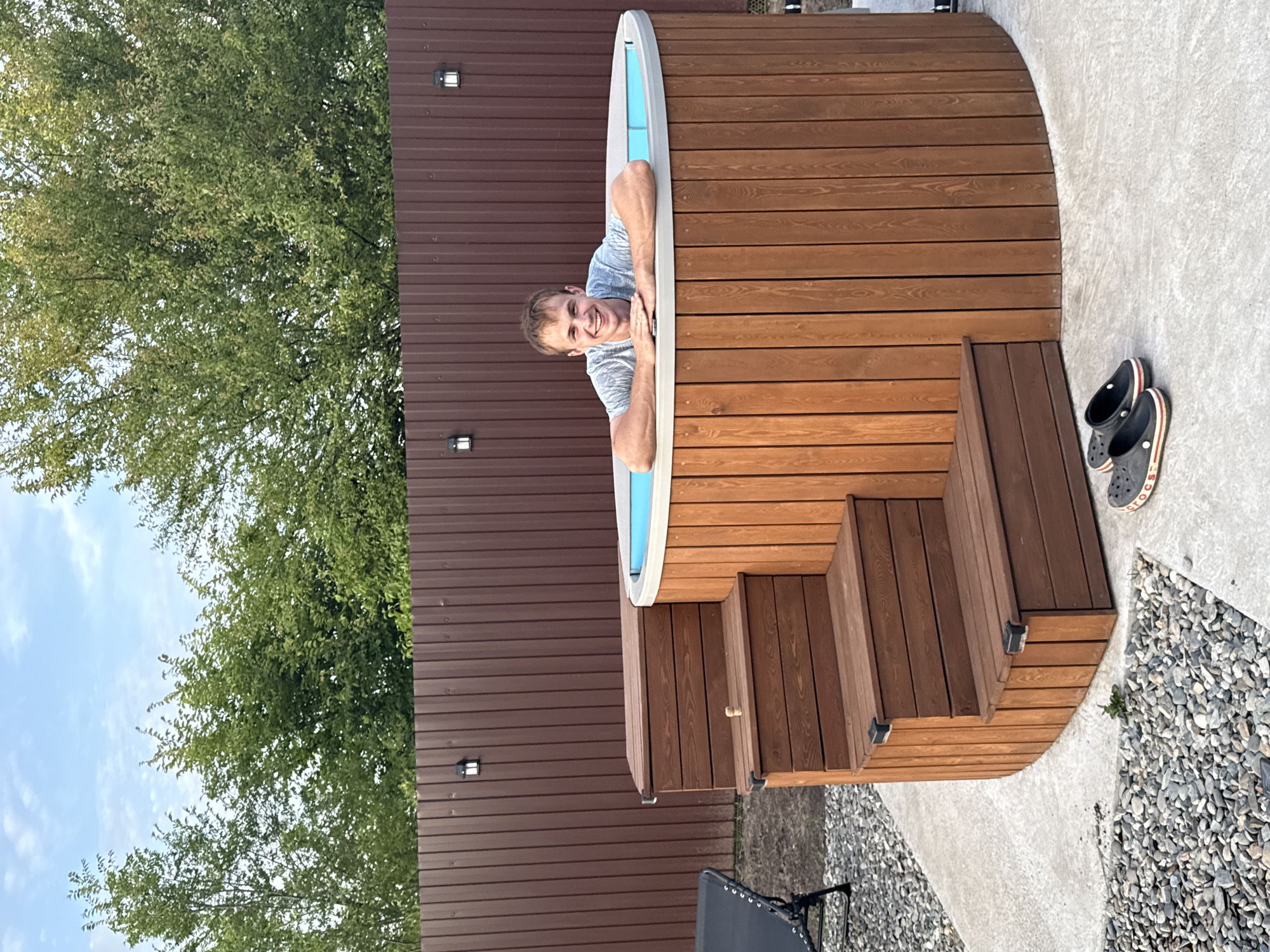 A man with short brown hair and a light blue and grey striped t-shirt smiles as he rests his arms on the edge of a wooden hot tub. He is leaning out of the tub, with his head and upper body visible. The hot tub is circular and made of wooden planks. Steps, also made of wood, lead up to one side of the hot tub. The scene appears to be an outdoor setting, with a brown metal fence in the background and lush green trees. The sky is visible, suggesting it is daytime with some clouds. A pair of black Crocs with red and white stripes are placed on the concrete ground next to the hot tub. There is no visible text in the image. The setting is likely a backyard or a recreational area in Starokorsunskaya, Russia. The man appears happy and relaxed.