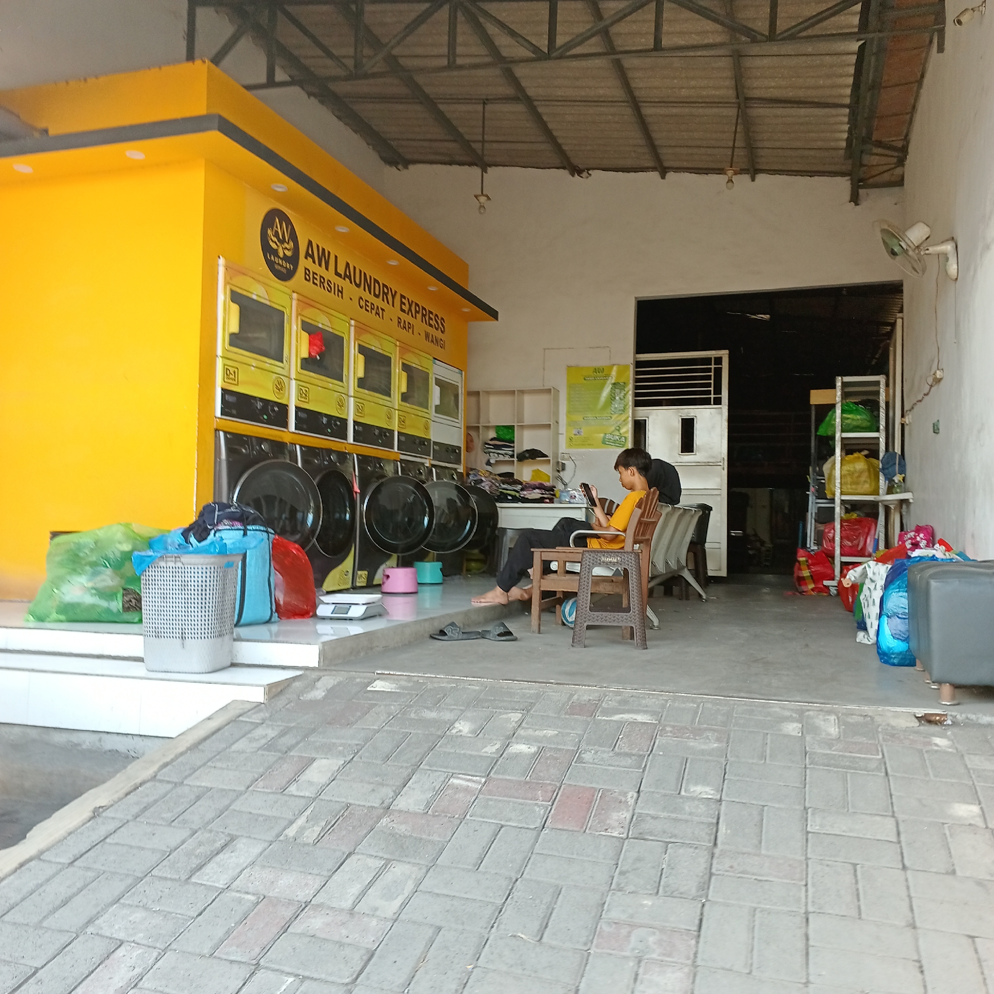 The image shows the exterior of a laundry service business, "AW LAUNDRY EXPRESS," located in Wedoro, Indonesia. The building features a bright yellow facade with the business name and slogan prominently displayed. Several commercial-grade washing machines are visible lined up along the front of the establishment. In front of the machines, laundry bags, a white laundry basket, and a weighing scale are placed on a white tiled platform. Two individuals are present in the scene: one person, wearing a yellow t-shirt and dark pants, is seated on a chair and looking at a mobile phone, while another person, in a black t-shirt, is partially visible behind them. The environment appears to be an open-air covered space, with a metal roof structure supported by beams. The foreground is a paved walkway made of interlocking bricks. Shelving units with various items, including laundry baskets and clothing, are visible inside the business premises. A wall-mounted fan is attached to the right wall.
