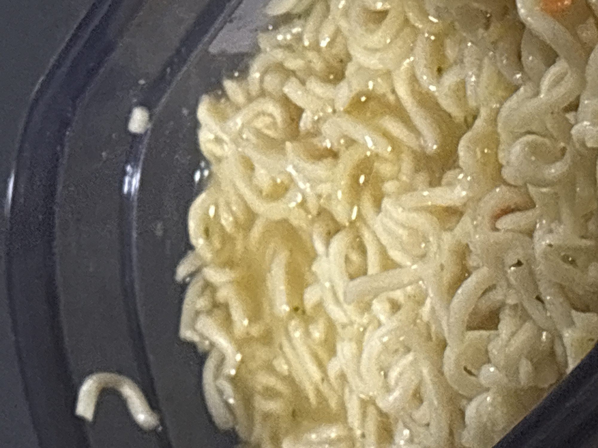 The image is a close-up, overhead shot of a bowl of instant ramen noodles. The noodles are pale yellow and appear to be cooked in a broth. There are some small, dark flecks visible in the noodles, possibly seasoning. The bowl itself is dark blue and appears to be made of plastic. The lighting is dim, and there is a slight glare on the surface of the broth. The background is out of focus and dark. There is no indication of a city or location.