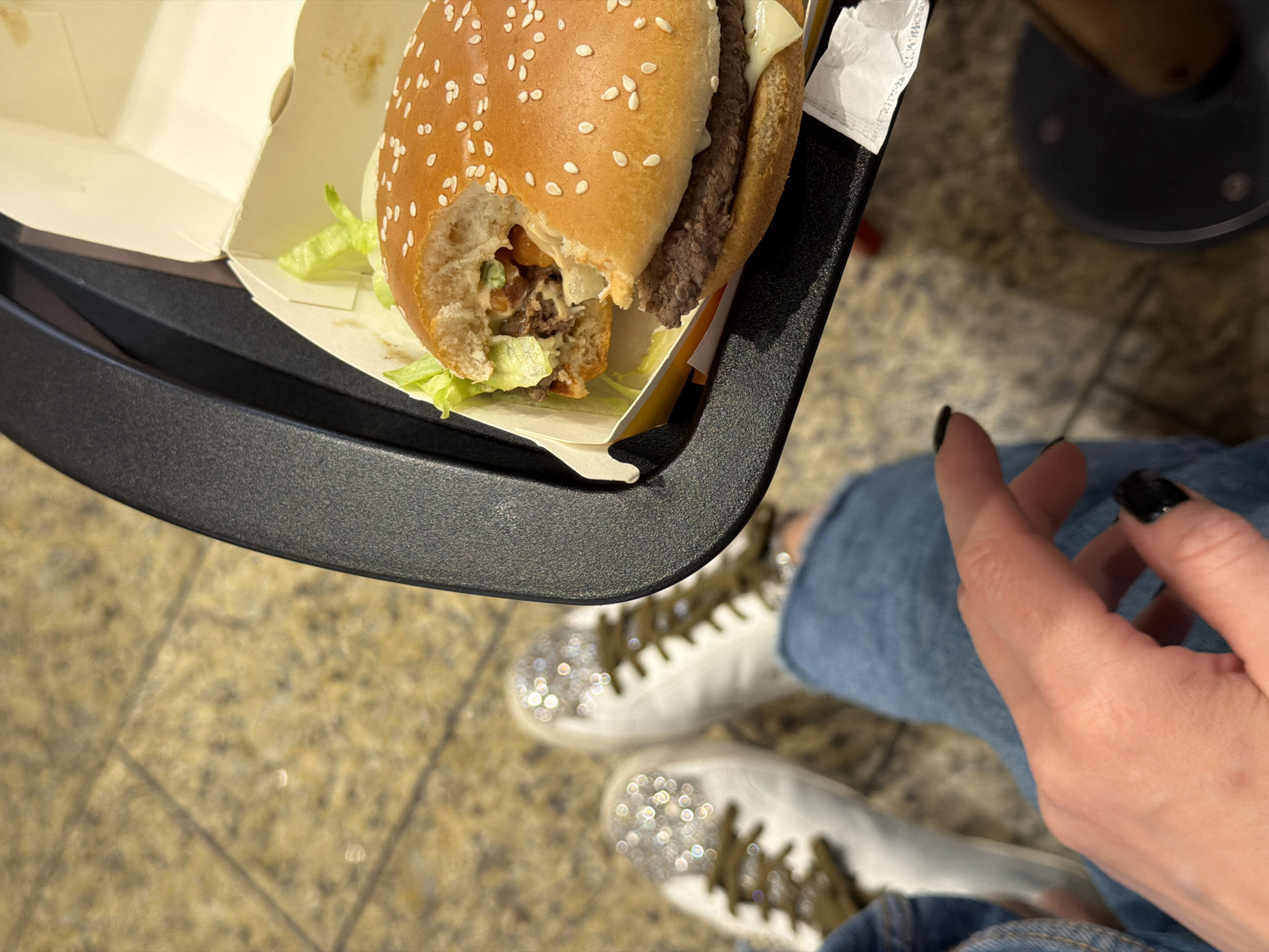 The image shows a partially eaten burger in an open box, held above a pair of legs clad in jeans. The feet are adorned with white sneakers featuring shimmering embellishments. The burger appears to be a fast-food type, containing lettuce, meat, and cheese, identifiable as a McDonald's burger. The burger and its box are resting on a dark gray tray. A hand with black nail polish is visible near the lower right corner. The background is a patterned floor, suggesting a public space, possibly a food court or restaurant. Based on the burger and the tray, the location is likely a McDonald's restaurant in Riga, Latvia.