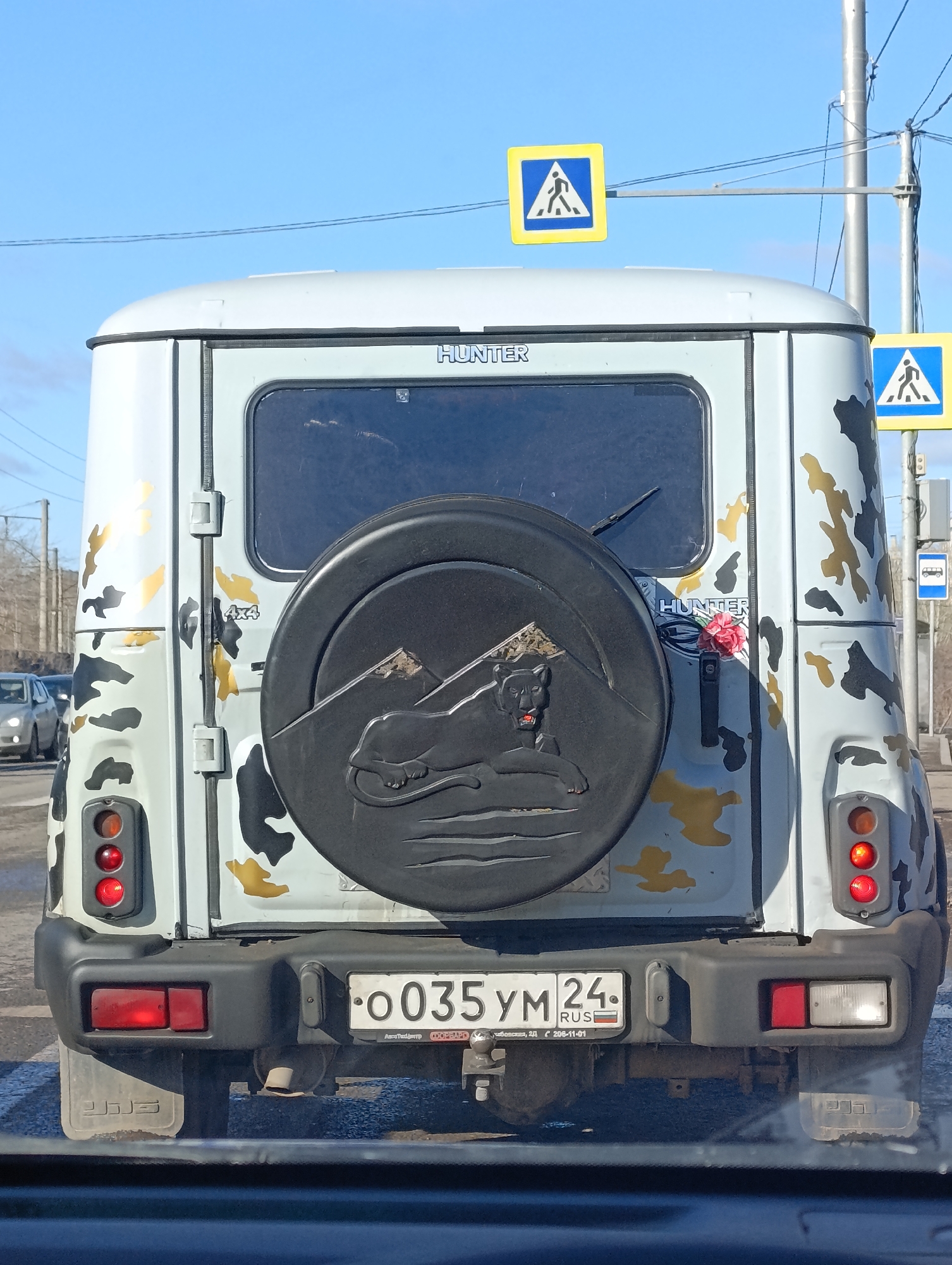 The image shows the rear of a light-gray UAZ Hunter SUV with camouflage patterning in black and tan, observed in Krasnoyarsk, Russia. The vehicle features a spare tire cover displaying an embossed mountain range and a reclining feline figure. The word "HUNTER" is printed on the upper and right sides of the rear door. A floral decoration is affixed near the "HUNTER" emblem. The license plate reads "O035YM24 RUS". Pedestrian crossing signs are visible above the vehicle. Other vehicles are faintly visible in the background. The perspective indicates the photo was taken from within another vehicle.