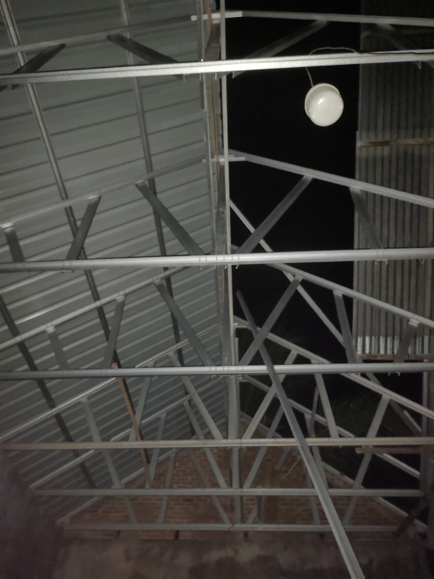 The image depicts the interior of a building under construction, specifically focusing on the roof structure and the corrugated metal roofing. The structure is a series of interconnected metal beams and supports, forming a triangular roof frame. The roofing material appears to be corrugated galvanized steel, laid out in parallel rows. A white, spherical light fixture hangs from one of the ceiling beams, casting a soft light on the surrounding structure. The background is dark, suggesting it is nighttime or the area is not yet illuminated. In the lower portion of the image, a brick wall is visible, indicating the building's walls are in place. The scene appears to be in Rayeukmatangkuli, Indonesia, a location known for its construction projects, particularly in the developing urban and rural areas.