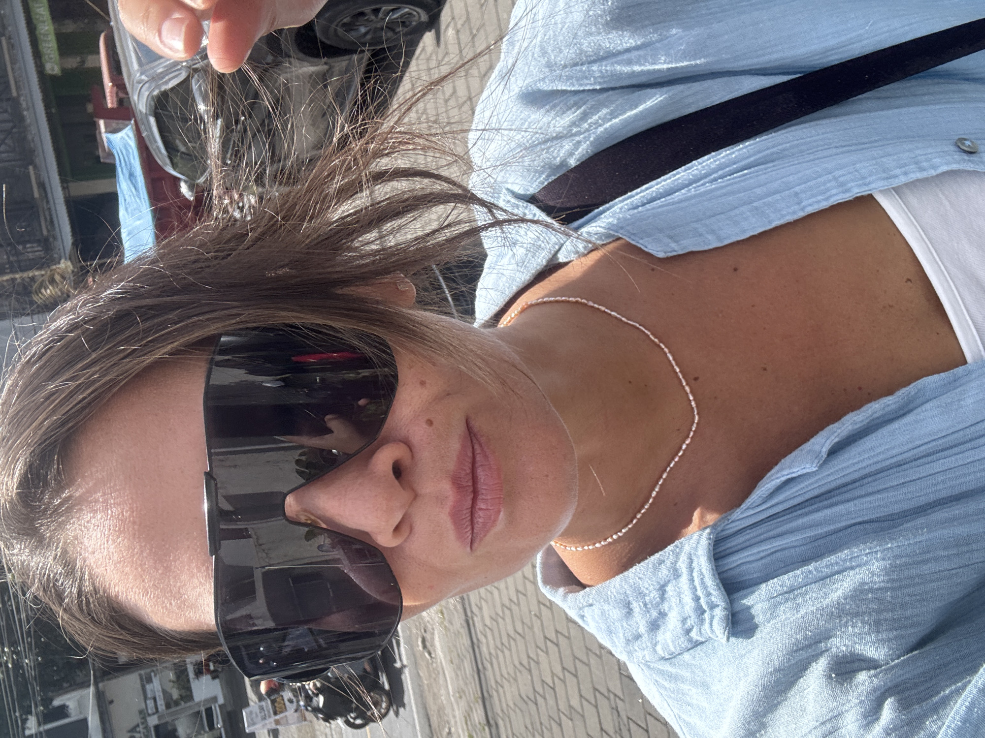 This is a close-up, angled selfie of a person with medium-toned skin, wearing large, dark sunglasses and a light blue, textured button-up shirt. A thin, beaded necklace is visible around their neck. The person's hair is brown and appears windblown. The background is blurred but suggests an outdoor setting with a street and what appears to be a car. A black strap, possibly from a bag, is visible across the person's chest. The lighting indicates it is daytime, likely sunny given the sunglasses. The clothing and sunny ambiance could suggest a warm climate. No visible text is present in the image.