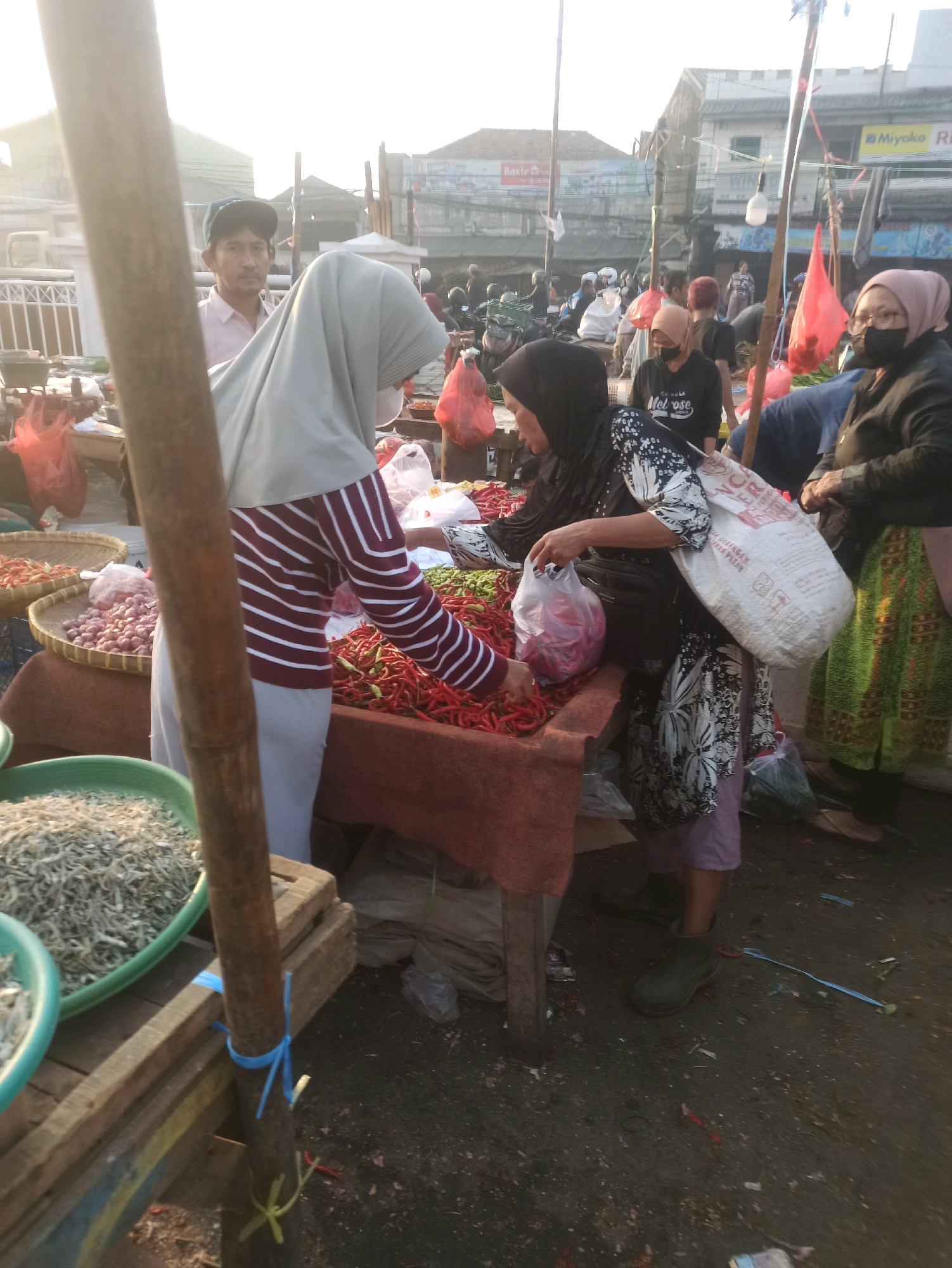 A street market in Cikarang, Indonesia, is captured in this medium shot, taken during daylight hours. The scene is bustling with activity as several individuals are engaged in commerce. In the foreground, a person wearing a light grey hijab and a striped maroon and white sweater stands behind a table laden with red chili peppers. To their right, another person, also in a hijab and a dark floral patterned dress, is placing chili peppers into a plastic bag. A large white woven bag is slung over their shoulder.
Behind the chili peppers, there are baskets filled with what appear to be shallots. To the left of the frame, a large green basin contains a pile of small dried fish. A wooden post, likely part of a market stall, stands prominently in the left foreground, secured with blue and yellow ties.
In the background, other market-goers are visible, some wearing masks. Buildings line the street, and several motorcycles are parked. The ground is dirt and appears somewhat littered. The overall impression is a typical scene of a vibrant Indonesian wet market.