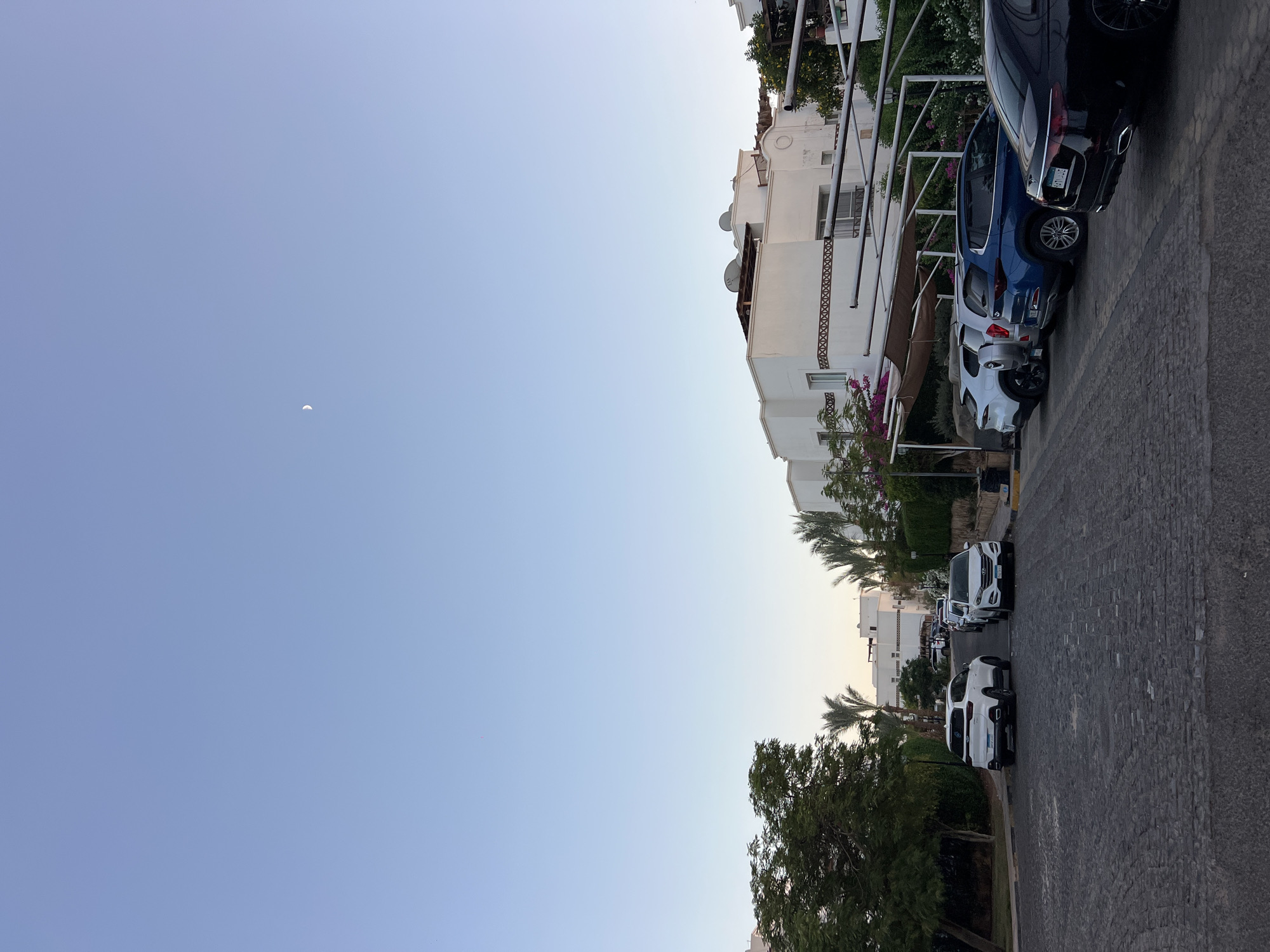The image depicts a paved street lined with parked cars in what appears to be a residential area or resort in Sharm El Sheikh, Egypt. The architecture of the buildings visible suggests a Mediterranean or North African style, characterized by white facades and arched details. A crescent moon is visible in the clear, pale blue sky, indicating it is likely early morning or late evening. Lush green trees and palm trees add to the tropical ambiance. The street itself is paved with dark, irregular cobblestones, and several vehicles, including SUVs and sedans, are parked along the side. The overall impression is one of a quiet, well-maintained urban or resort setting.