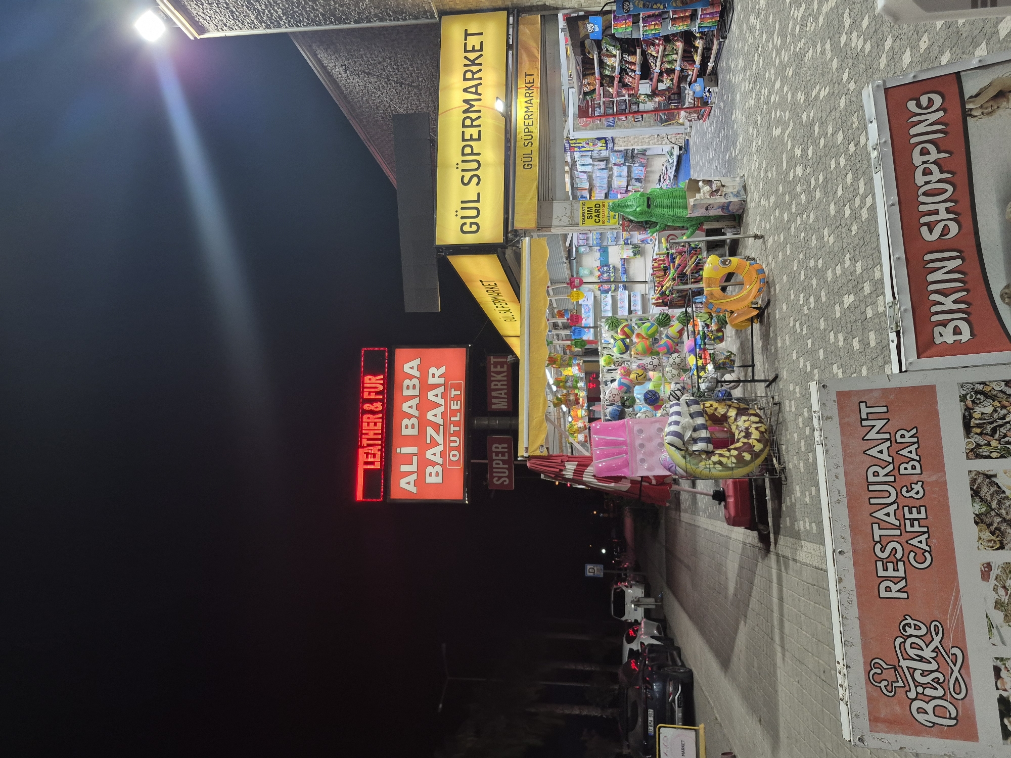 The image depicts a nighttime scene in Manavgat, Turkey, likely along a commercial street. A prominent red neon sign reads "LEATHER & FUR ALI BABA BAZAAR OUTLET." To the right, a bright yellow sign announces "GÜL SUPERMARKET," with another smaller "GÜL SUPERMARKET" sign below it. The entrance to the supermarket is visible, with shelves stocked with goods. In front of the supermarket, a display of colorful toys, inflatable items, and novelty items, including a green dinosaur figurine, is set up. Several cars are parked on the street in the background. In the foreground, two signs are visible: one advertising "BISTRO RESTAURANT CAFE & BAR" with images of food, and another proclaiming "BIKINI SHOPPING" with a partial image of a woman. The ground is paved with interlocking grey tiles. The overall atmosphere suggests a busy tourist area with various retail offerings.