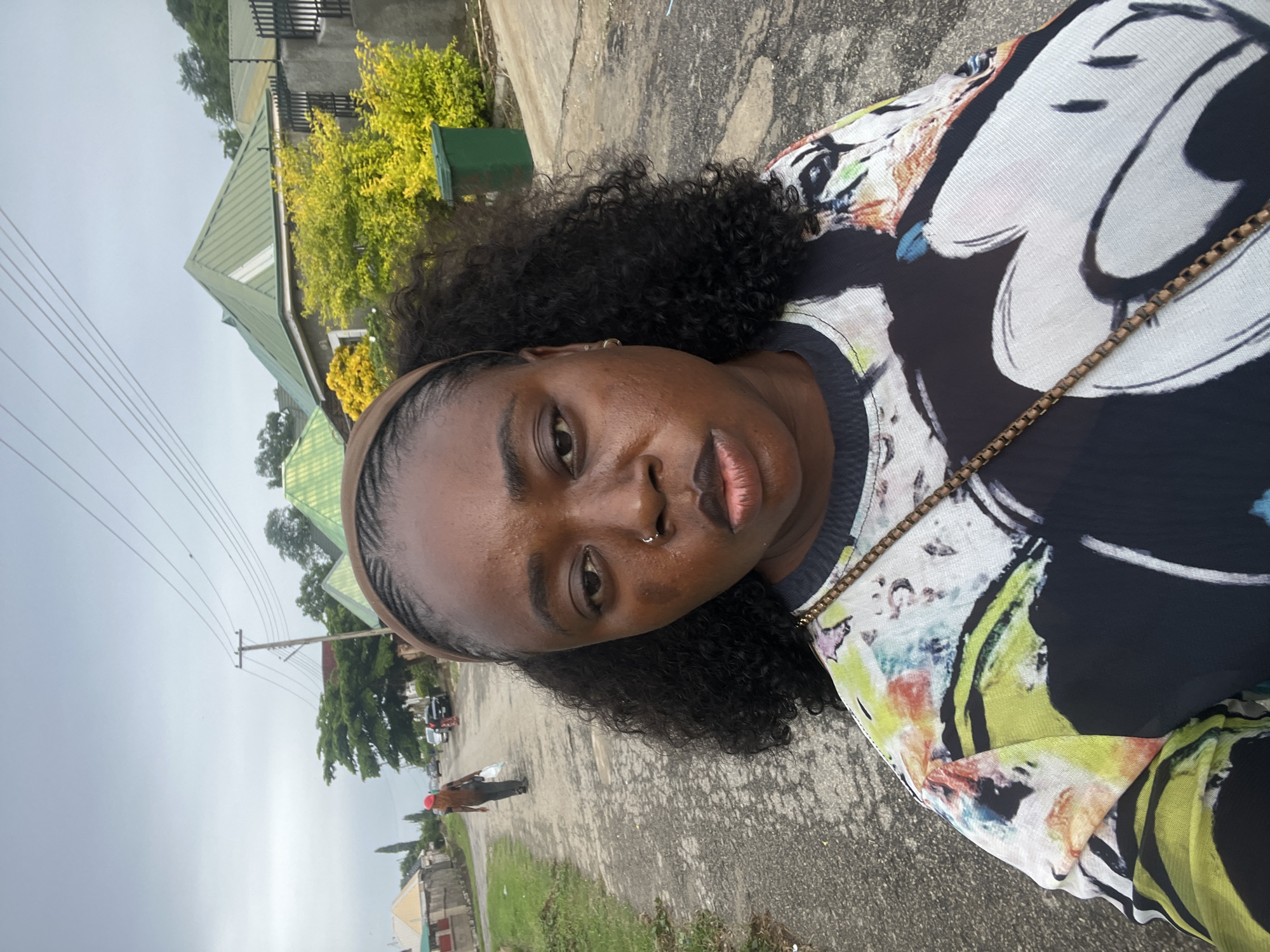 In Abuja, Nigeria, an outdoor, medium shot captures a woman in a close-up selfie against a slightly overcast sky and a street scene. The woman, positioned centrally, has dark skin, curly dark hair tied back with a brown headband, and wears a patterned shirt featuring large black and white Mickey Mouse imagery with colorful abstract designs. She has a nose ring and appears to be looking directly at the camera with a neutral expression.
Behind her, a paved street stretches into the distance. A person wearing a red cap and carrying a white bag walks away from the camera down the street. Further back, several cars are parked on the side of the road. The background includes residential buildings with green, sloped roofs, some with grey stone walls and black metal fences. Lush green and yellow foliage, along with a green refuse bin, are visible near one of the houses. Utility poles and power lines are present overhead. The overall environment suggests a residential area in Abuja.