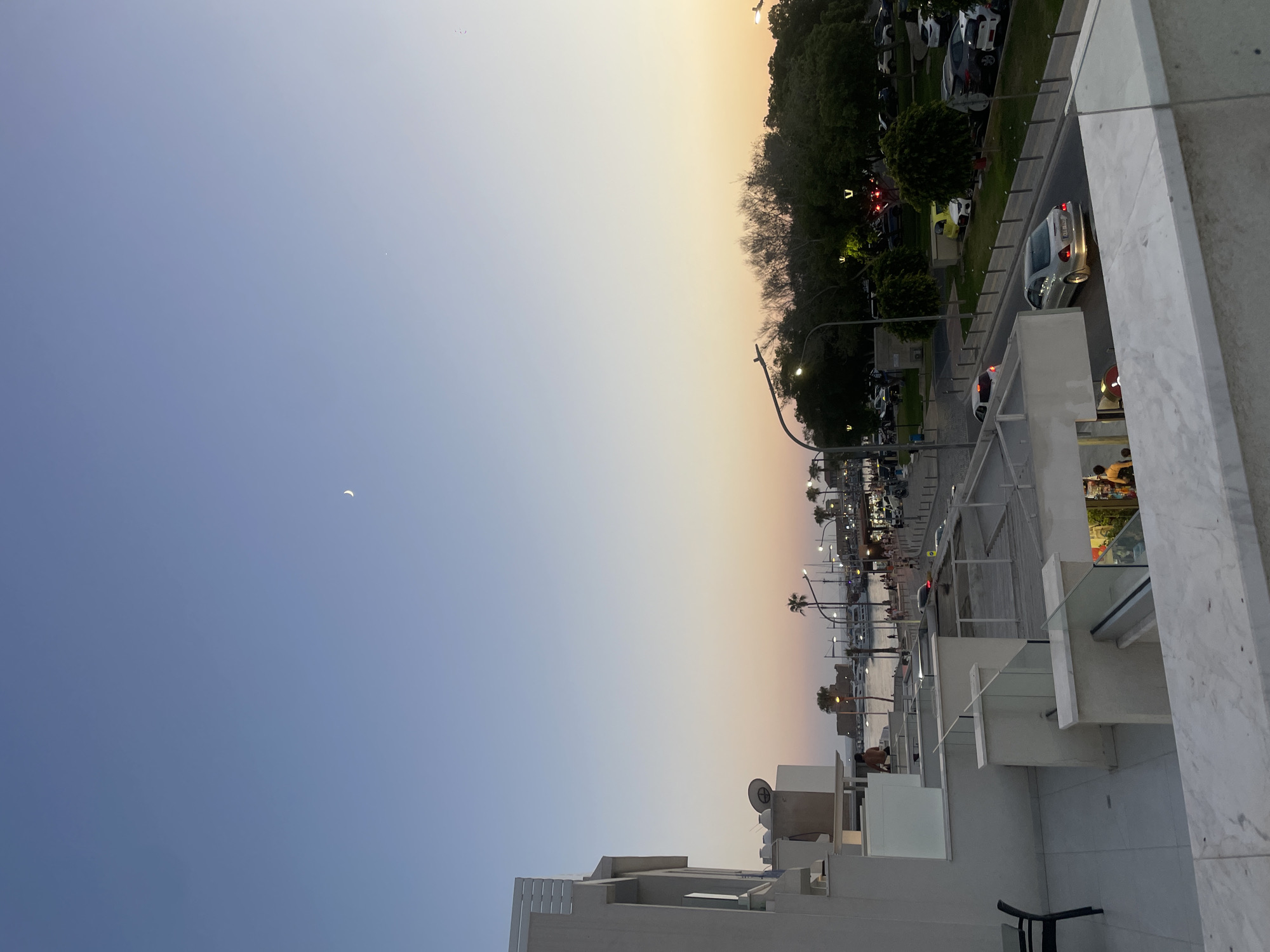 The image captures a view from a balcony overlooking a coastal area in Paphos Municipality, Cyprus, during twilight. In the foreground, a modern building with white balconies and glass railings is visible. A person is seated on a chair on the balcony. Below, a street is lined with parked cars and streetlights. Further in the distance, a harbor is filled with boats, and a historic castle is illuminated by lights. Palm trees and lush green foliage border the street and harbor area. The sky is a gradient of pale blue and soft orange, with a crescent moon faintly visible. The overall scene depicts a serene evening by the sea in a European coastal town.