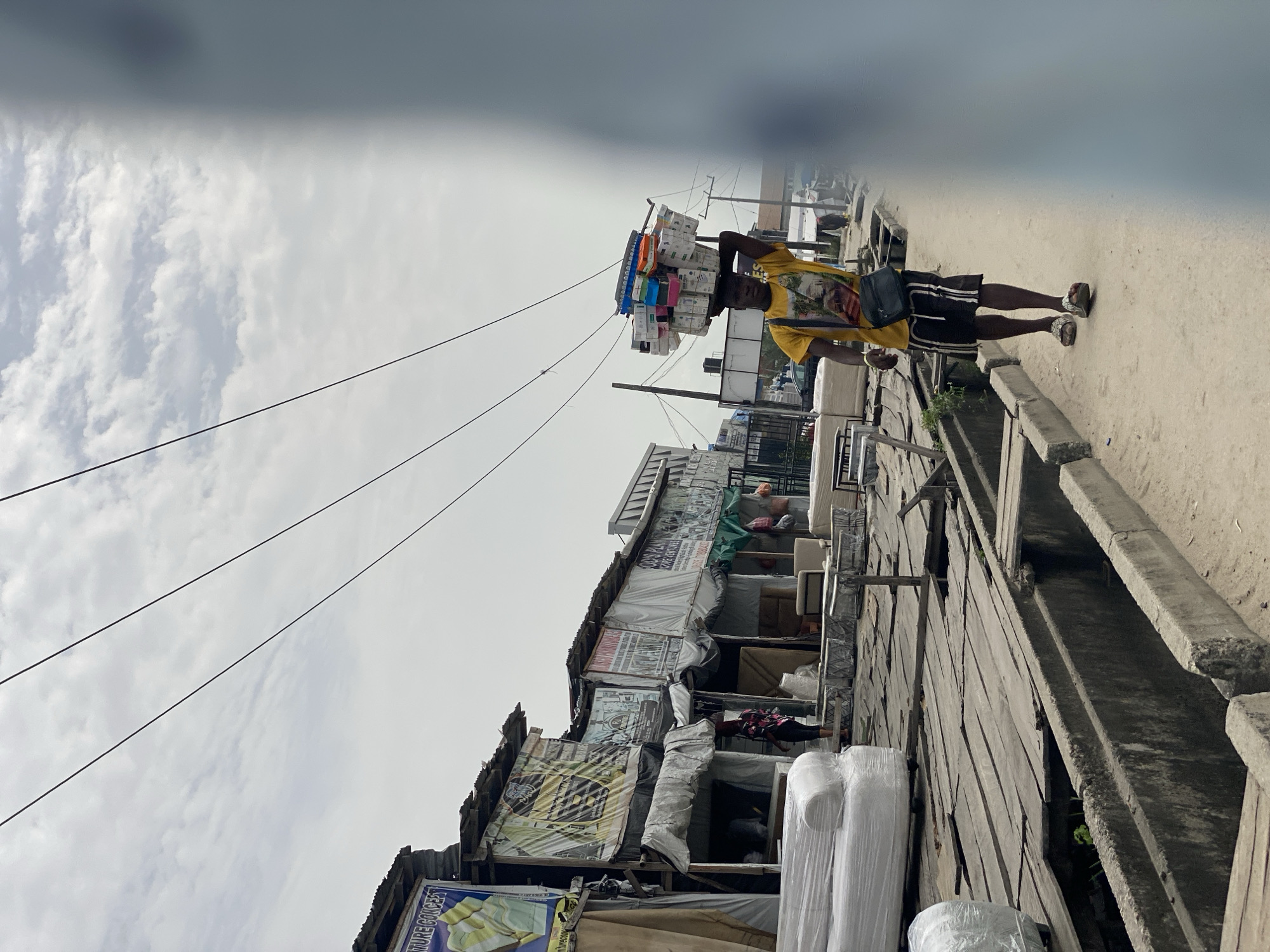 A street scene in Ado, Nigeria, depicts a man walking on a raised concrete walkway next to a road. He is carrying a large stack of items on his head, and is wearing a yellow t-shirt and black shorts. A black bag is slung over his shoulder. To his left are buildings, some appearing to be shops, with signs and posters attached to the exterior. Some furniture, potentially for sale, is visible. Overhead are multiple electrical or communication wires. The sky is overcast.