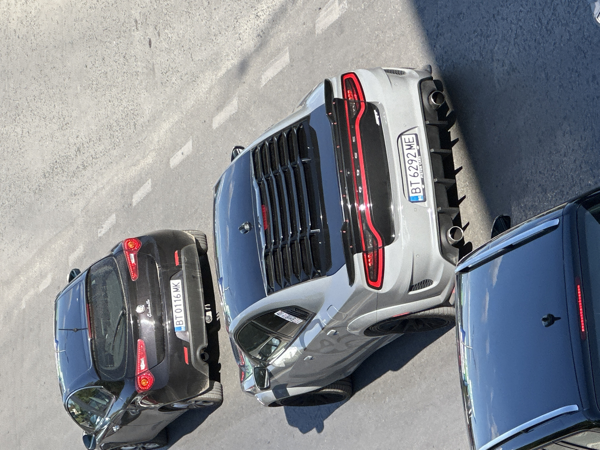 In Veliko Tarnovo, Bulgaria, on a paved road with white lane markings, three cars are visible from an elevated perspective. The uppermost car is a black Alfa Romeo Giulietta hatchback with license plate "BT 0116 MK". Directly below it is a silver Dodge Charger SRT Hellcat with a black roof and a distinctive rear spoiler, its license plate reads "BT 6292 ME". In the foreground, the roof of a black car, possibly an SUV, with silver roof rails is visible. The scene is illuminated by daylight, casting shadows on the road.