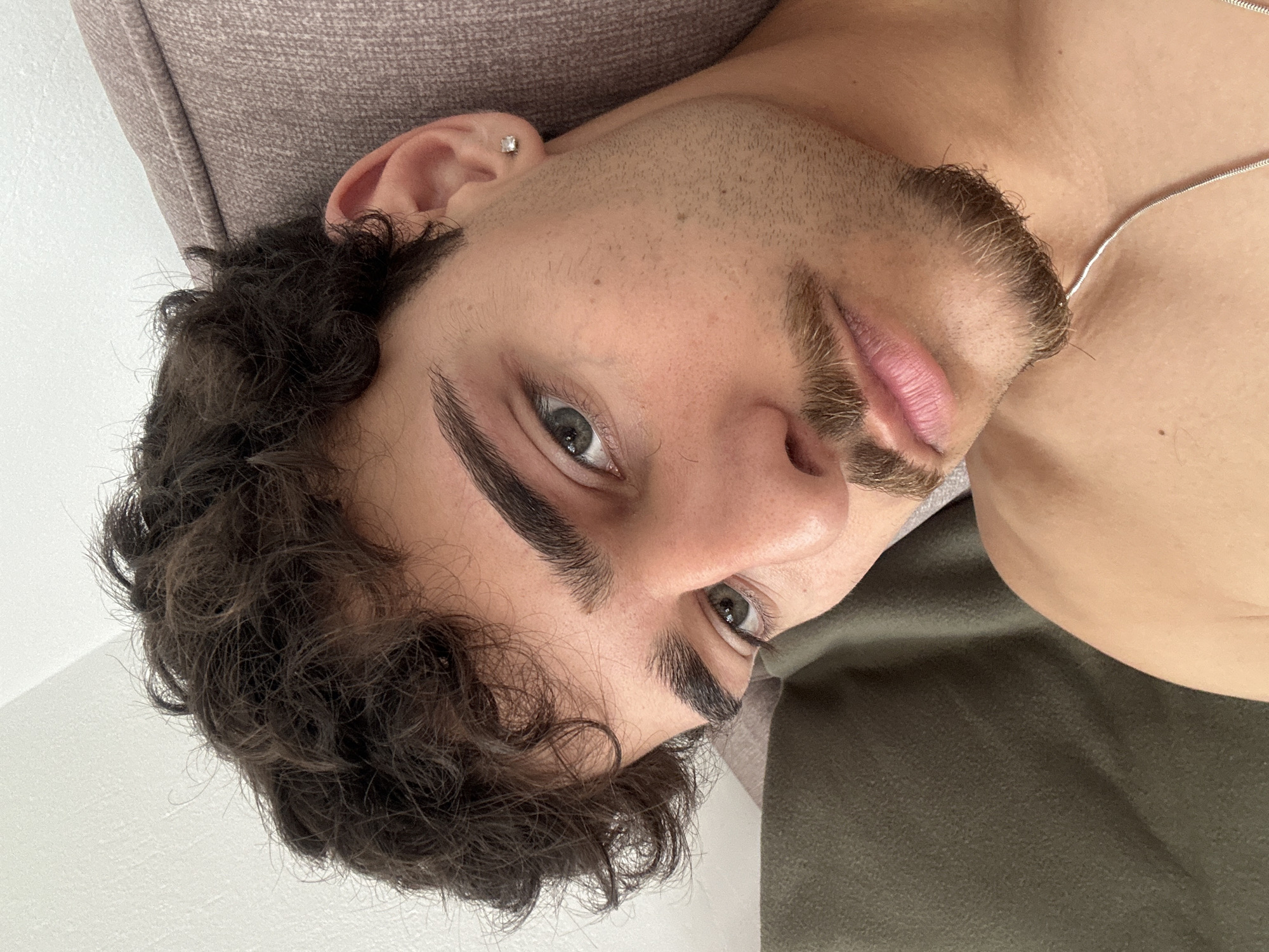 A close-up, slightly downward-angled shot of a light-skinned male with dark curly hair, a short beard, and light-colored eyes. He is lying on a light brown surface, likely a cushion or pillow, with a dark olive green fabric visible in the background. He appears to be wearing a silver necklace and has a small stud earring in his left ear. The image does not provide any information about the location being Troyan, Bulgaria.
