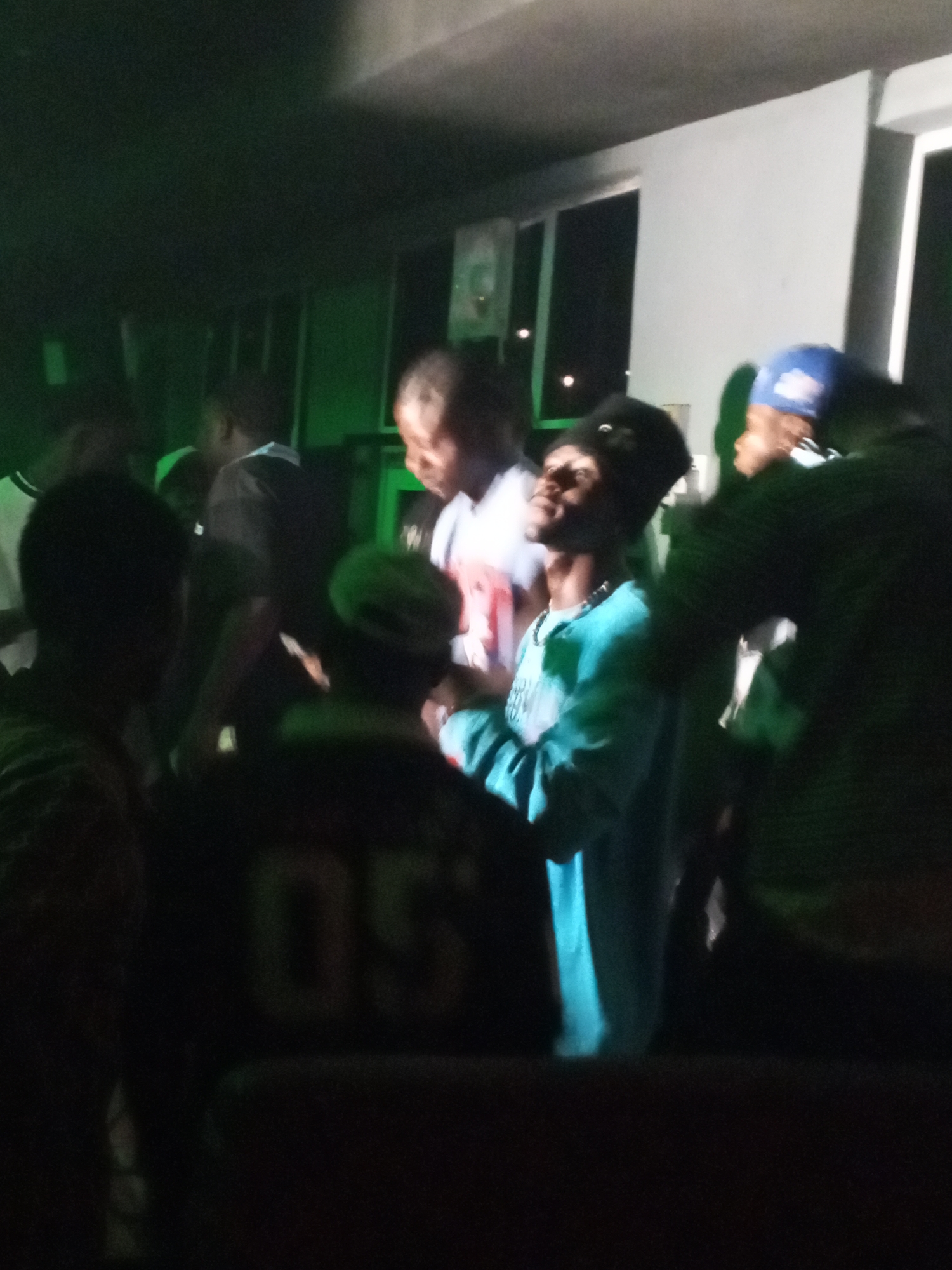 The image shows a group of people indoors, likely in Lokoja, Nigeria, based on the implied cultural context. The lighting is dim, with a green cast in some areas. Several individuals are visible, including a man in a blue long-sleeved shirt looking upward, and another wearing a blue baseball cap. A person with a garment displaying "05" is partially visible from the back. The setting appears to be near a window or a lit exterior, suggested by the brighter areas behind the group.